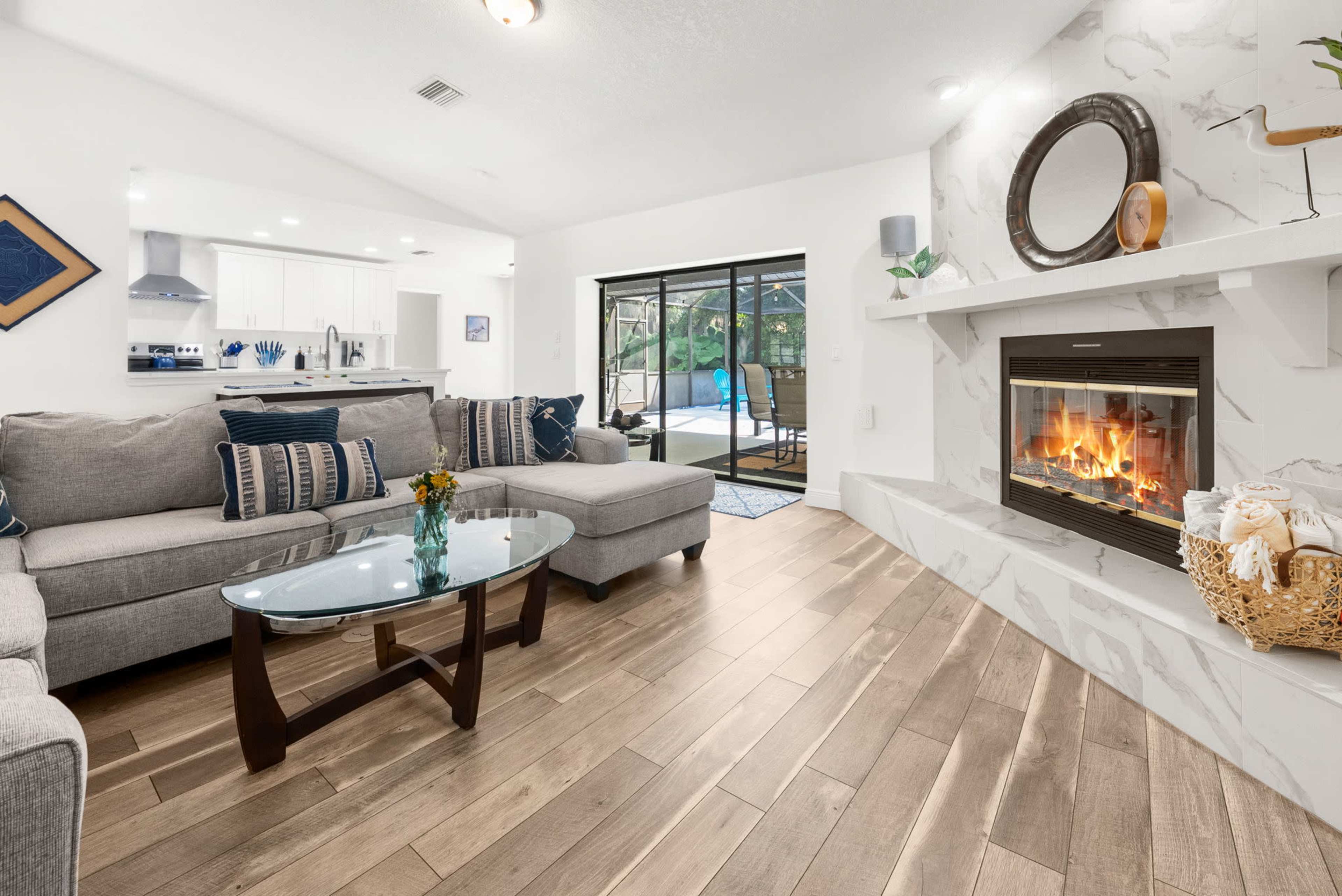 A modern living room features a gray sectional sofa, a glass coffee table, and a fireplace with a marble surround, overlooking a patio area through sliding glass doors.