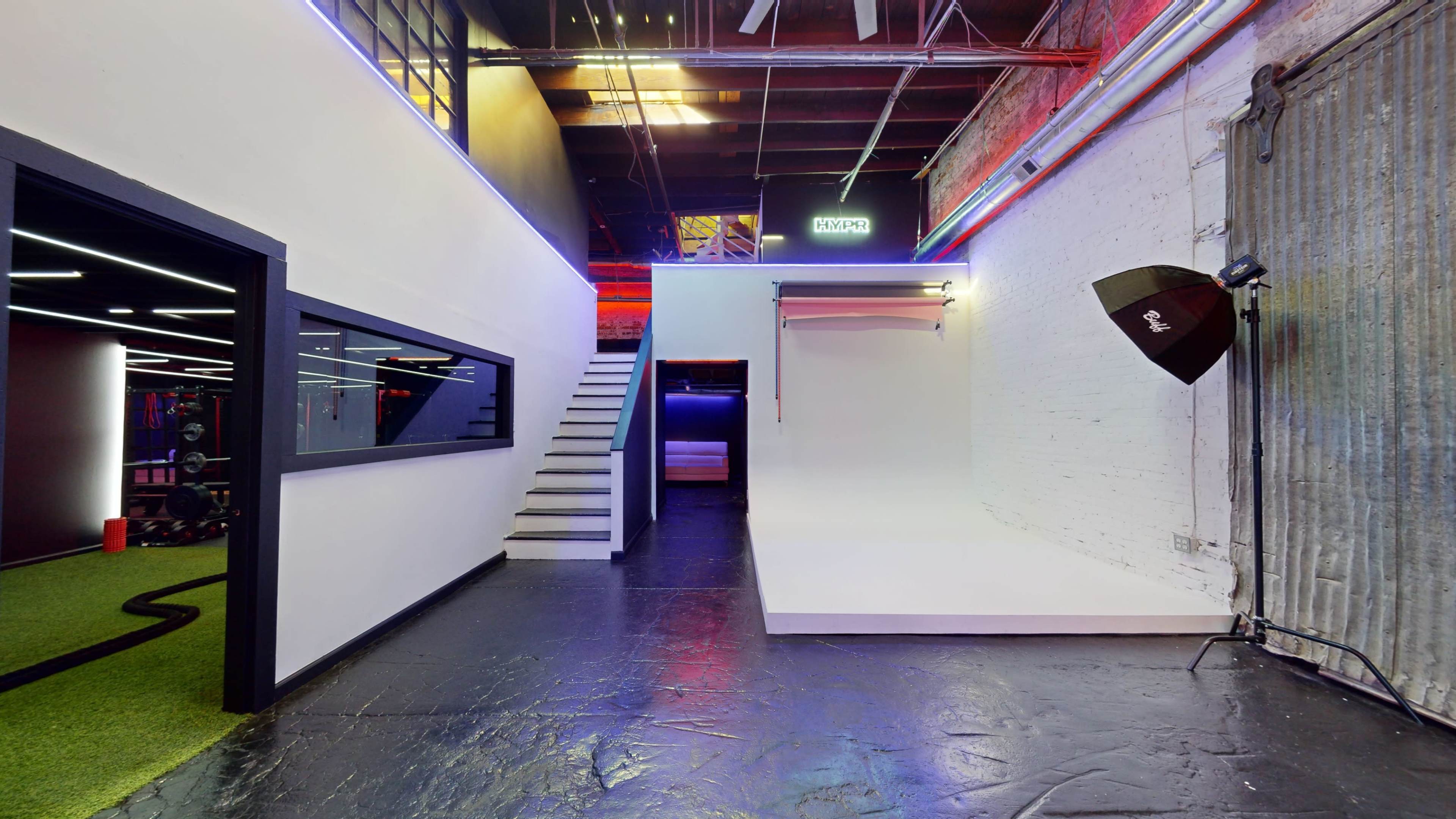 An industrial-style interior featuring a staircase leading to an upper level, a white backdrop against a wall, and studio lighting equipment.