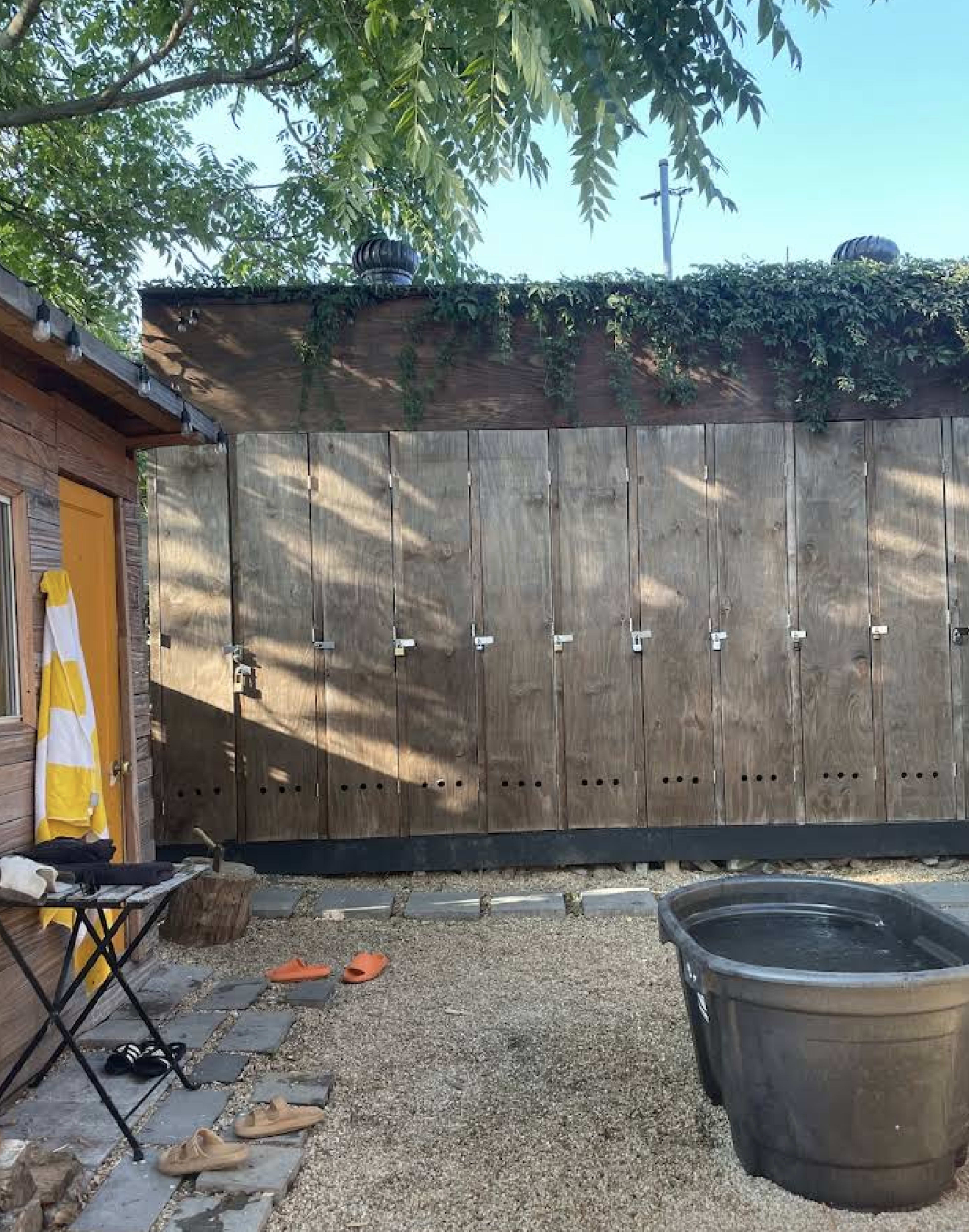 The image shows a row of wooden lockers against a wall, with a black tub and a few personal items on the ground in a gravel area.