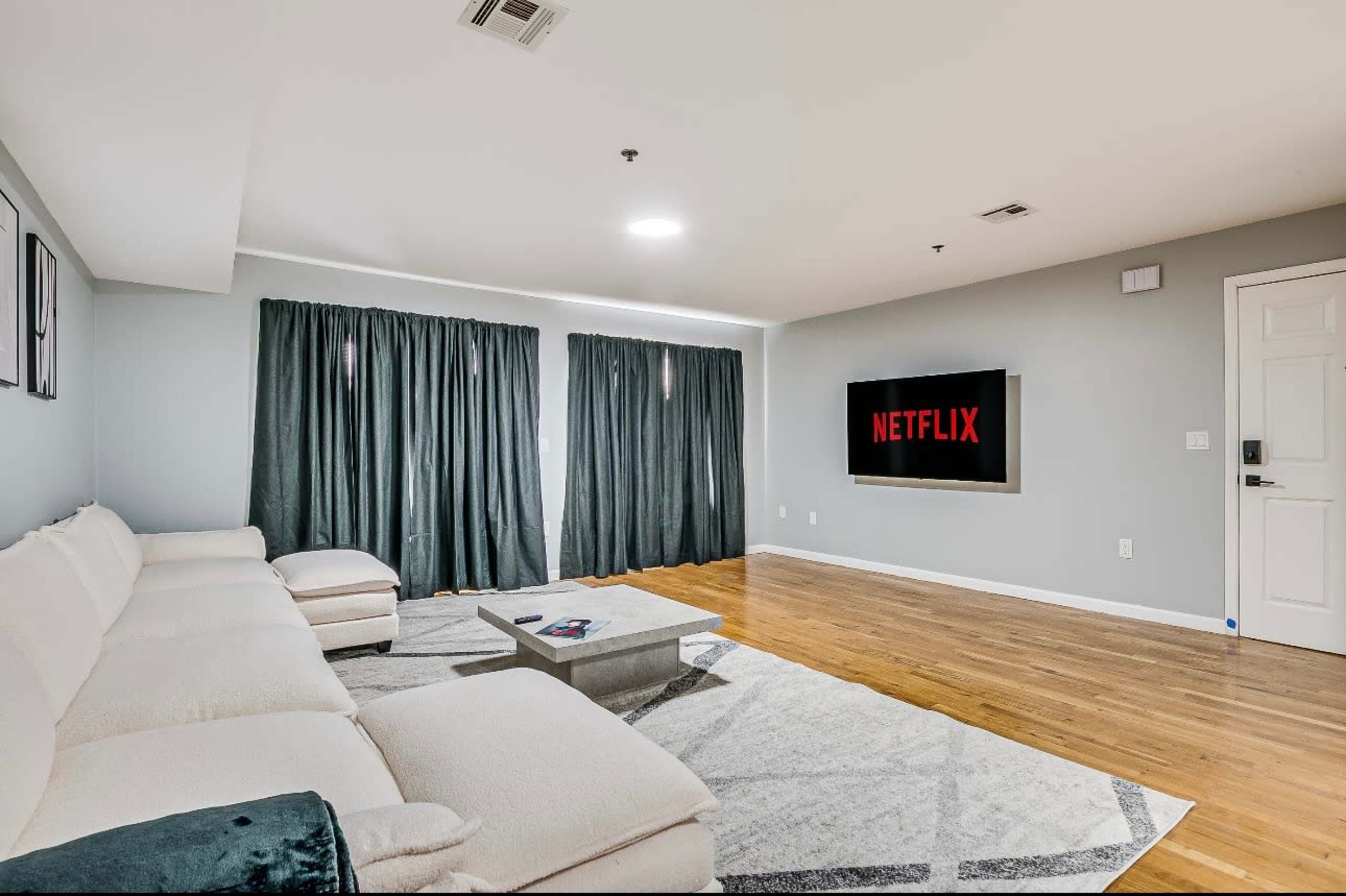 The living room features a white sofa, a gray rug, and a television displaying the Netflix logo, with dark curtains covering the windows.