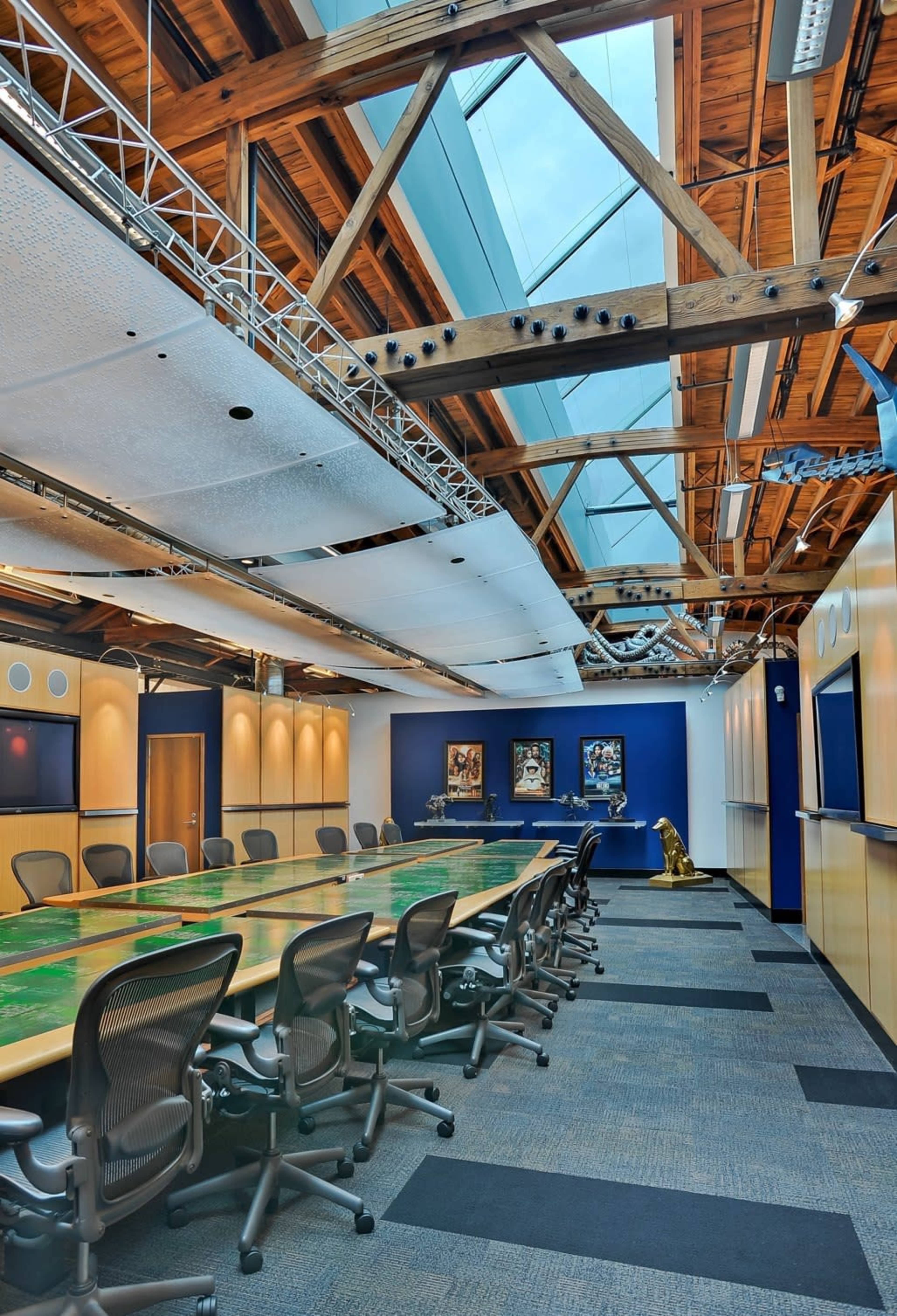 A spacious conference room features a long wooden table surrounded by ergonomic chairs, with exposed wooden beams and a skylight above.