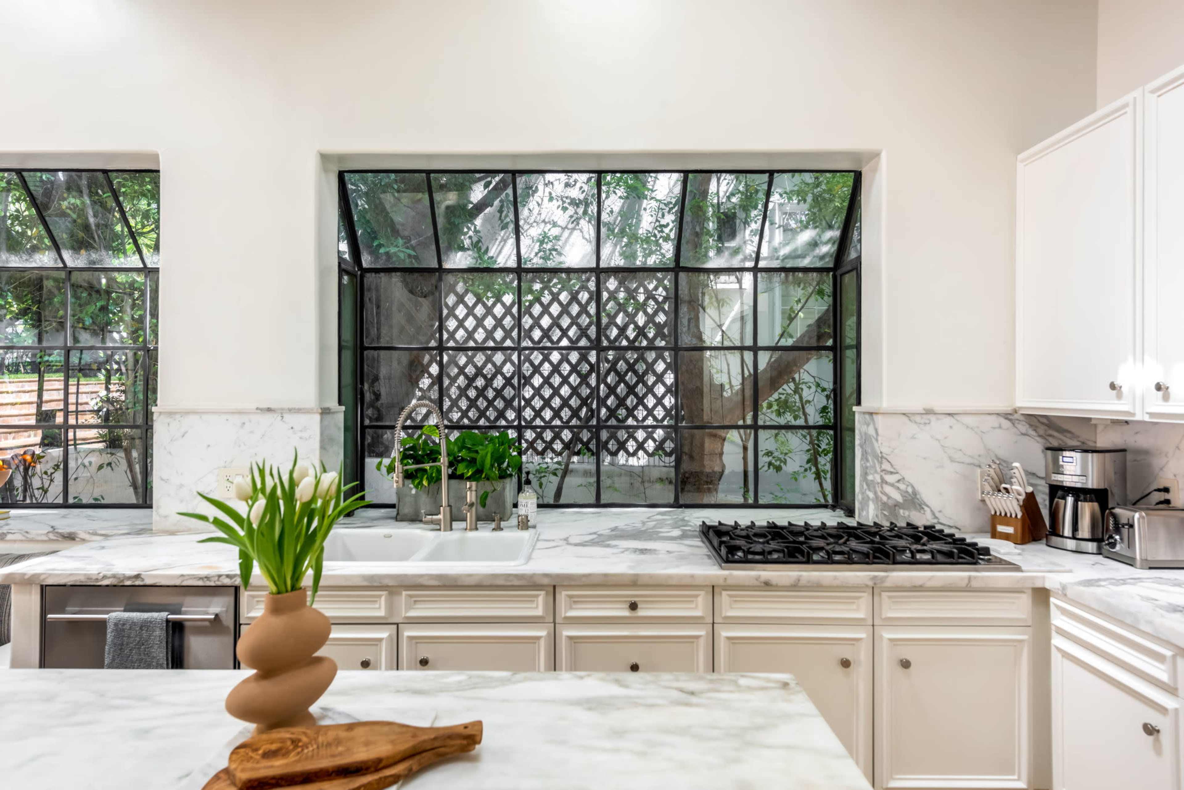 A modern kitchen features white cabinetry, a marble countertop, and large windows that provide a view of a garden space.