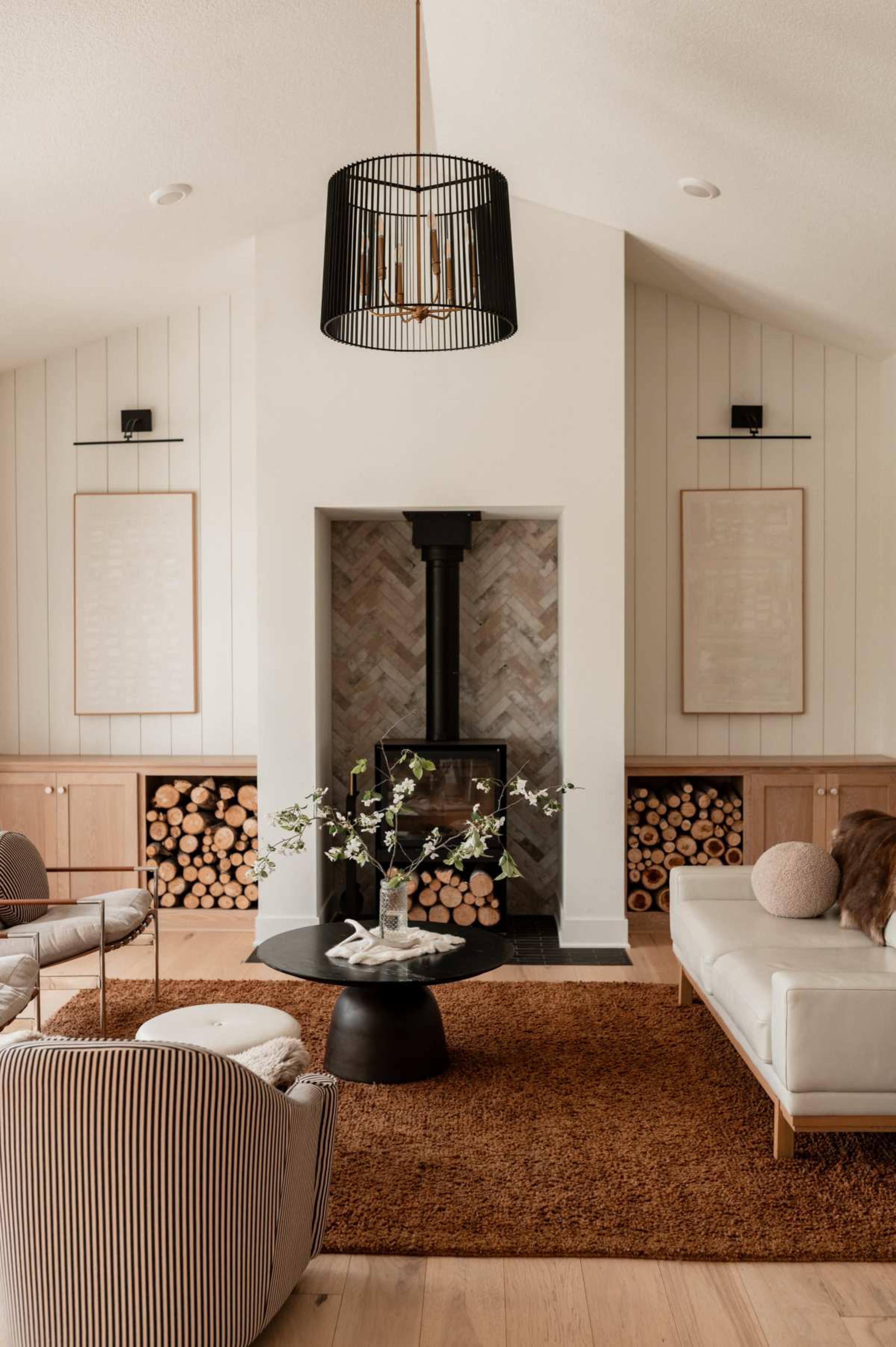 A modern living room features a black pendant light, a fireplace with herringbone tiles, and neatly stacked firewood on either side, complemented by a round coffee table and minimalist furniture.