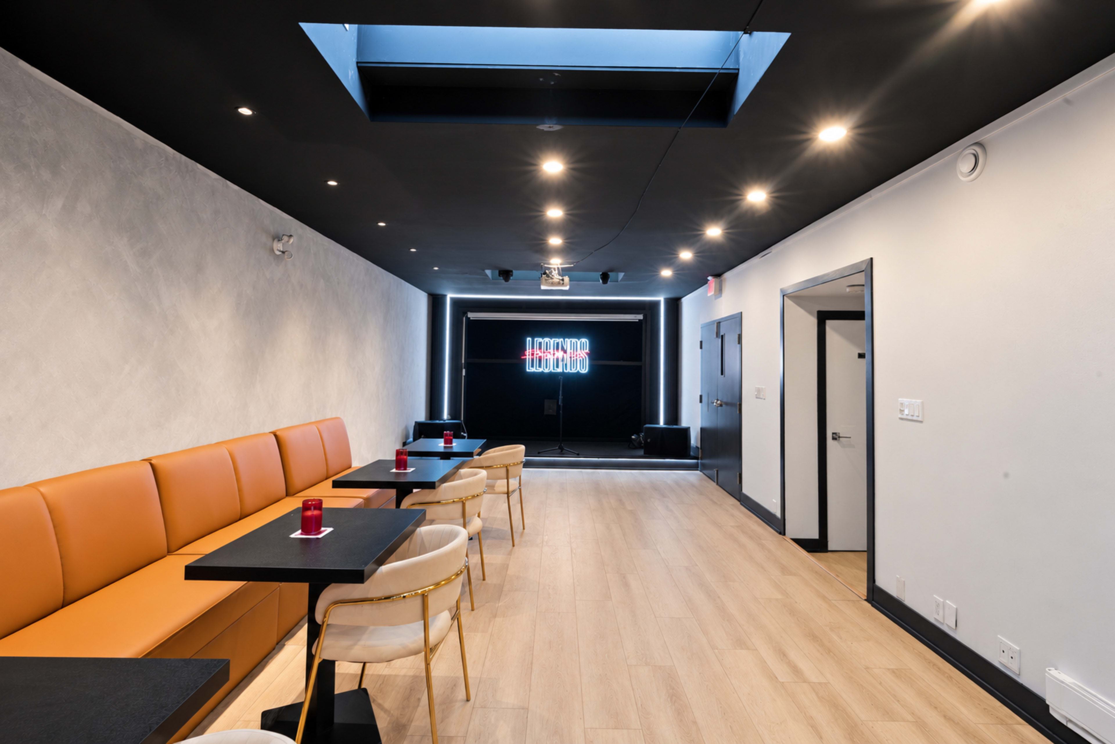 The image shows a modern, well-lit interior of a restaurant or lounge with an orange banquette, black tables, and a projector screen at the far end.