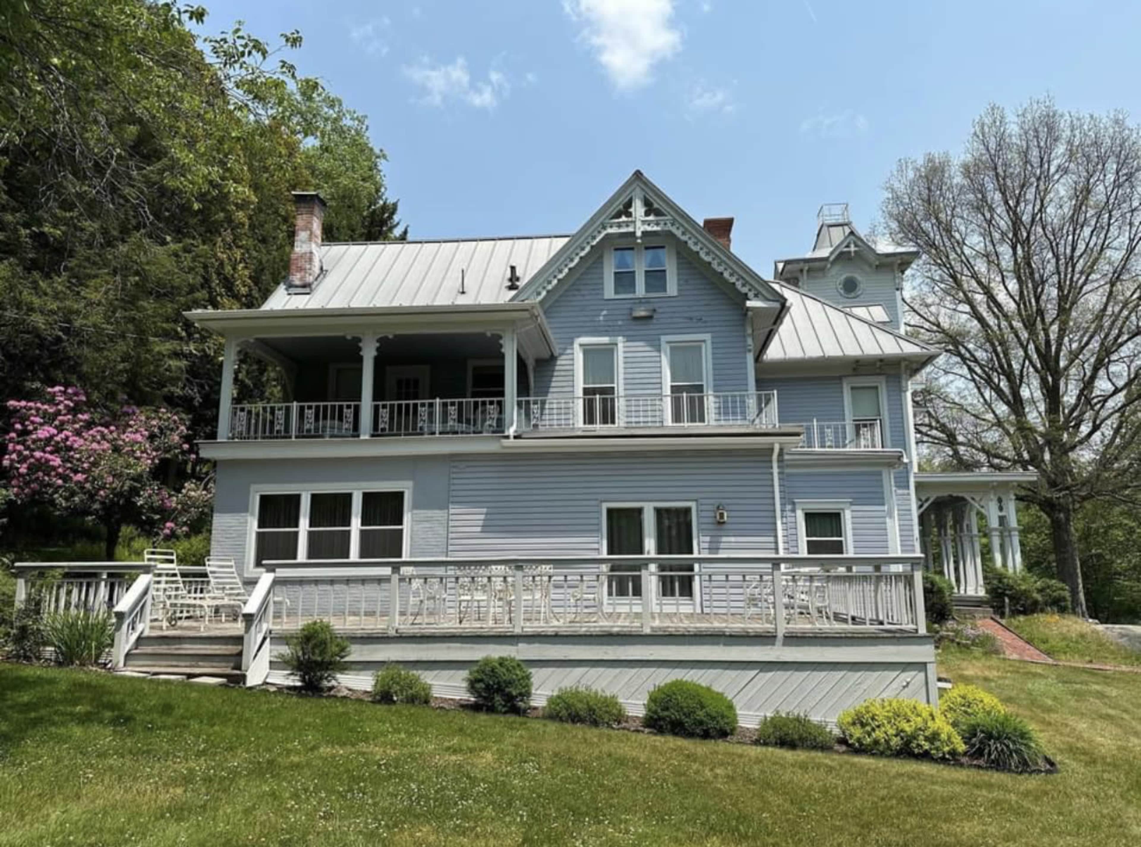 A large, two-story house with a light blue exterior and a wrap-around porch surrounded by greenery.