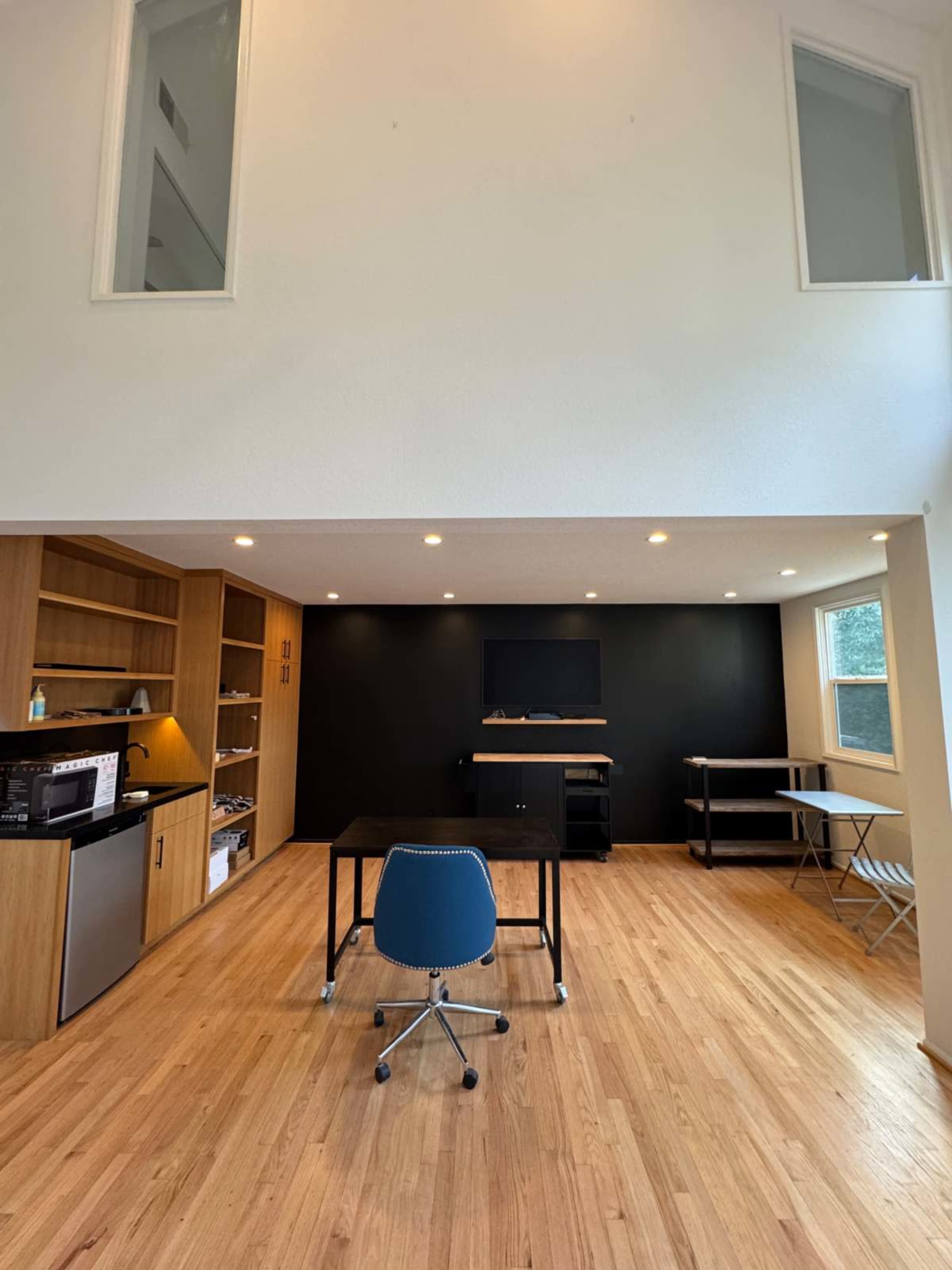 The image shows a modern interior workspace featuring a wooden floor, a black accent wall with a mounted TV, and a desk with a chair, surrounded by shelves and a mini refrigerator.