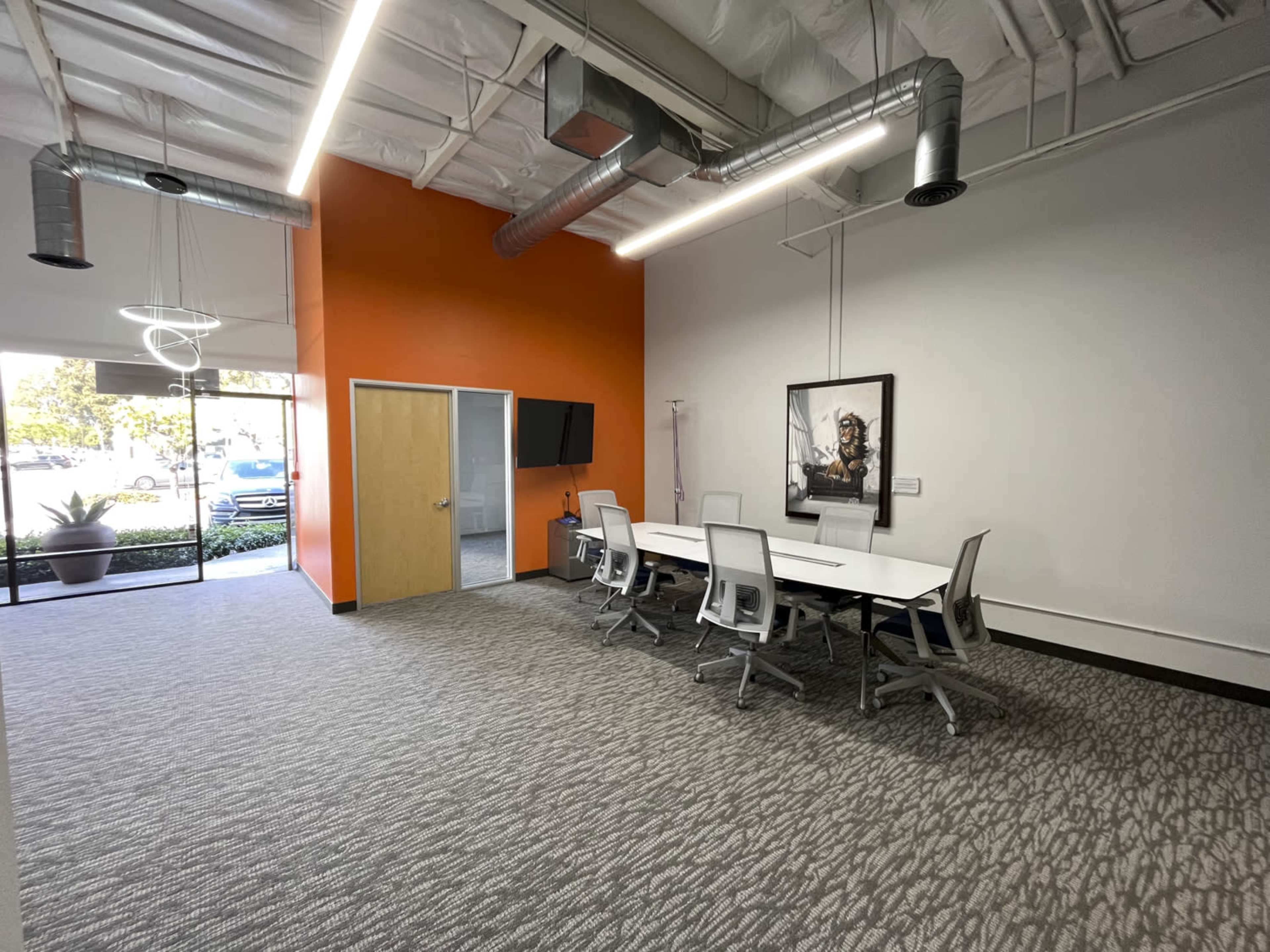 A modern conference room with a long white table, ergonomic chairs, and an orange accent wall, alongside large windows providing natural light.