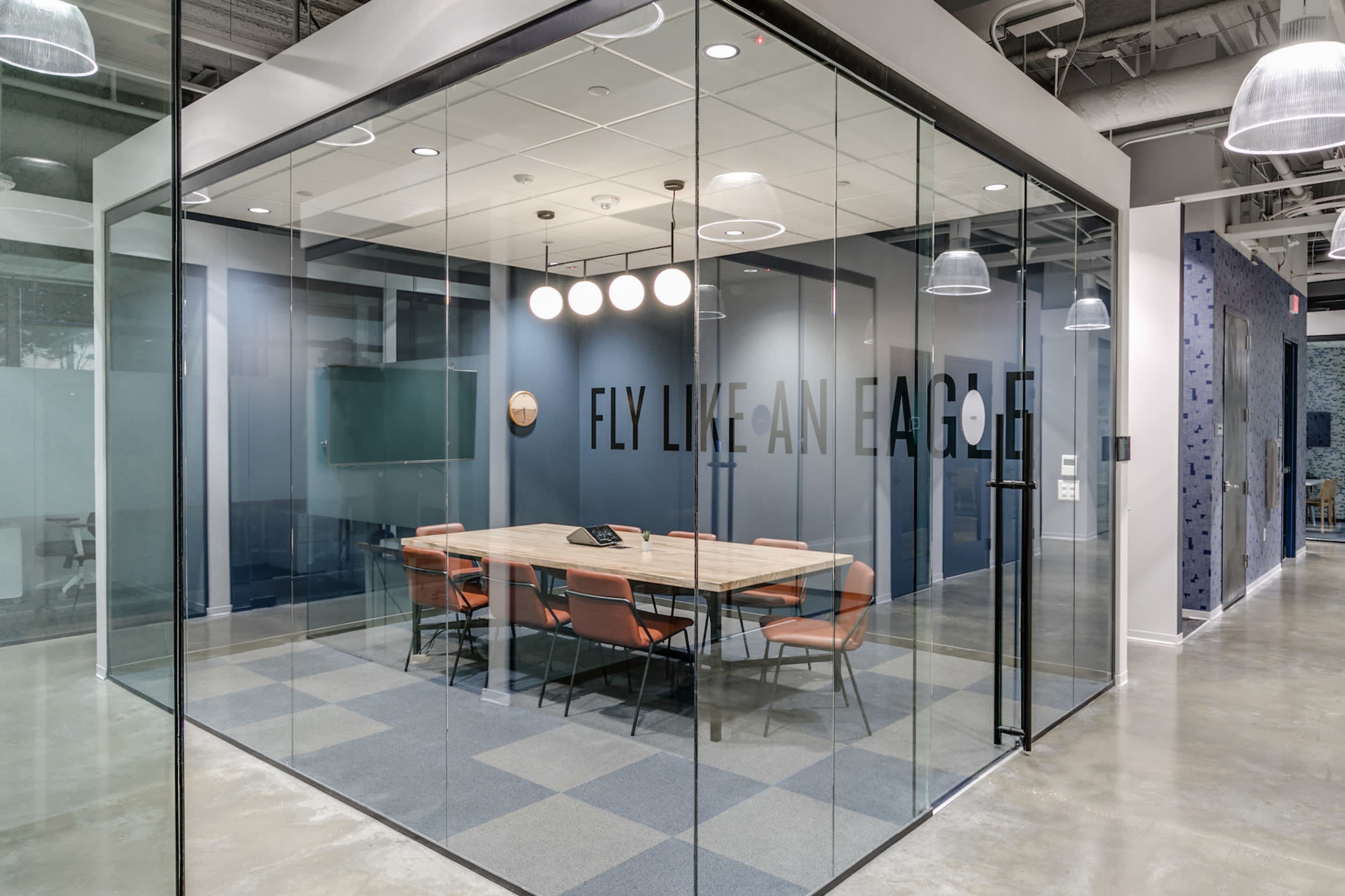 A modern conference room with a large table and orange chairs, enclosed by glass walls and featuring the phrase "FLY LIKE AN EAGLE" on one side.
