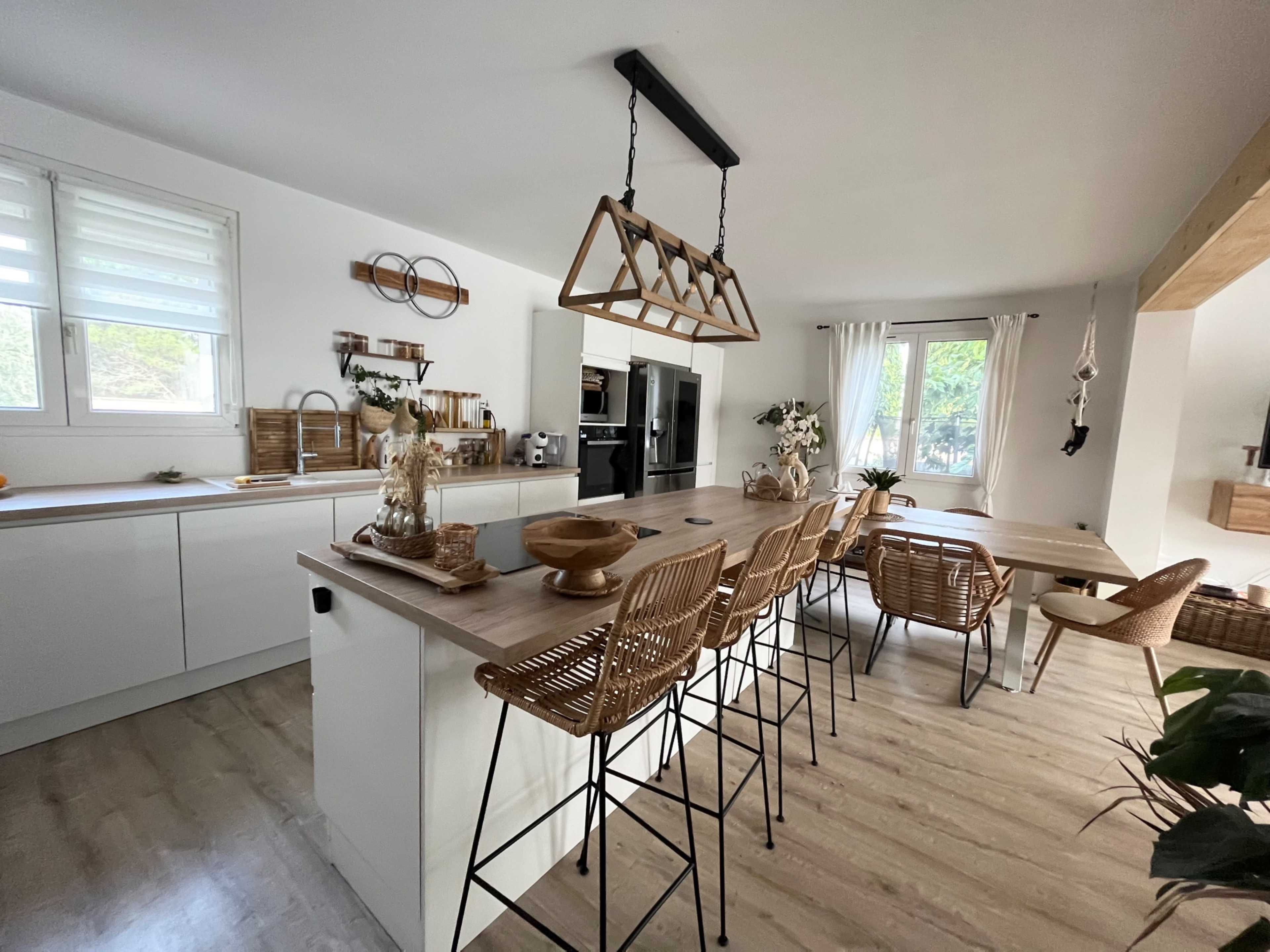 The image shows a modern kitchen with a central island, bar stools, and a dining table, featuring light-colored cabinetry and wooden accents.