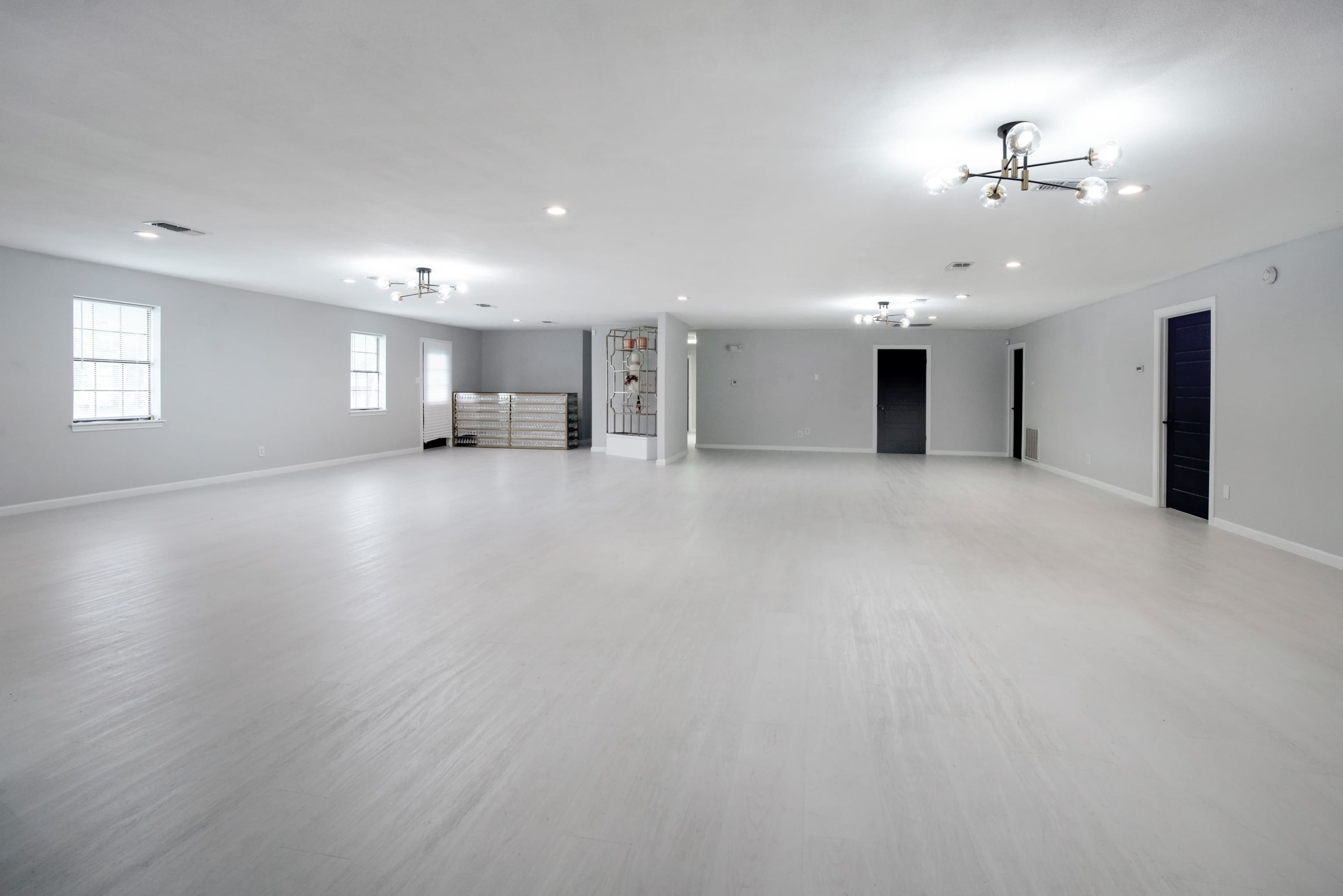 The image shows a spacious, empty room with white walls and flooring, featuring several light fixtures and two windows.