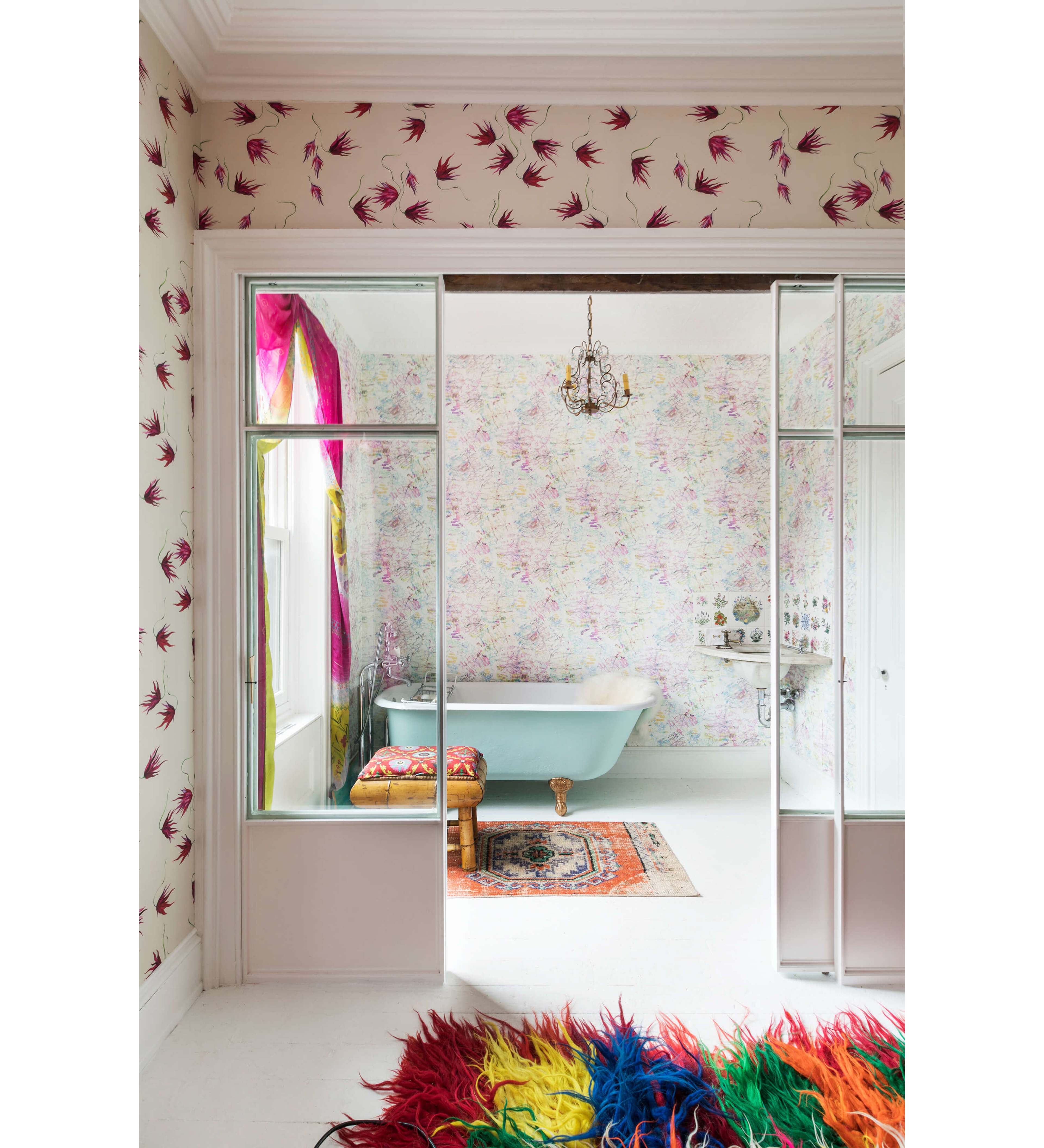 The image shows a brightly decorated bathroom featuring a freestanding bathtub, floral wallpaper, and colorful curtains, viewed through an open doorway.