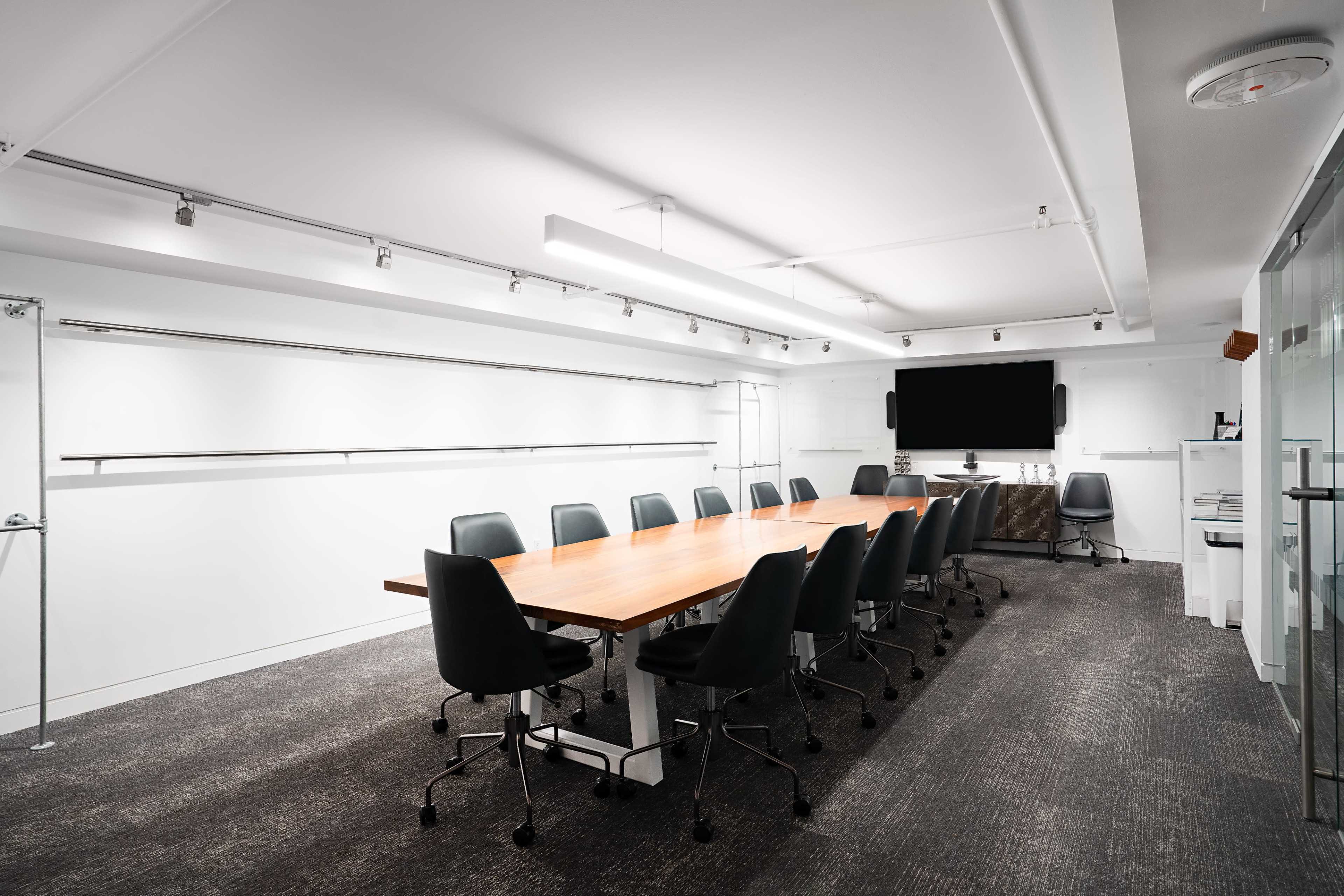 A modern conference room features a long wooden table surrounded by black chairs, with a large screen mounted on the wall and a minimalist design.