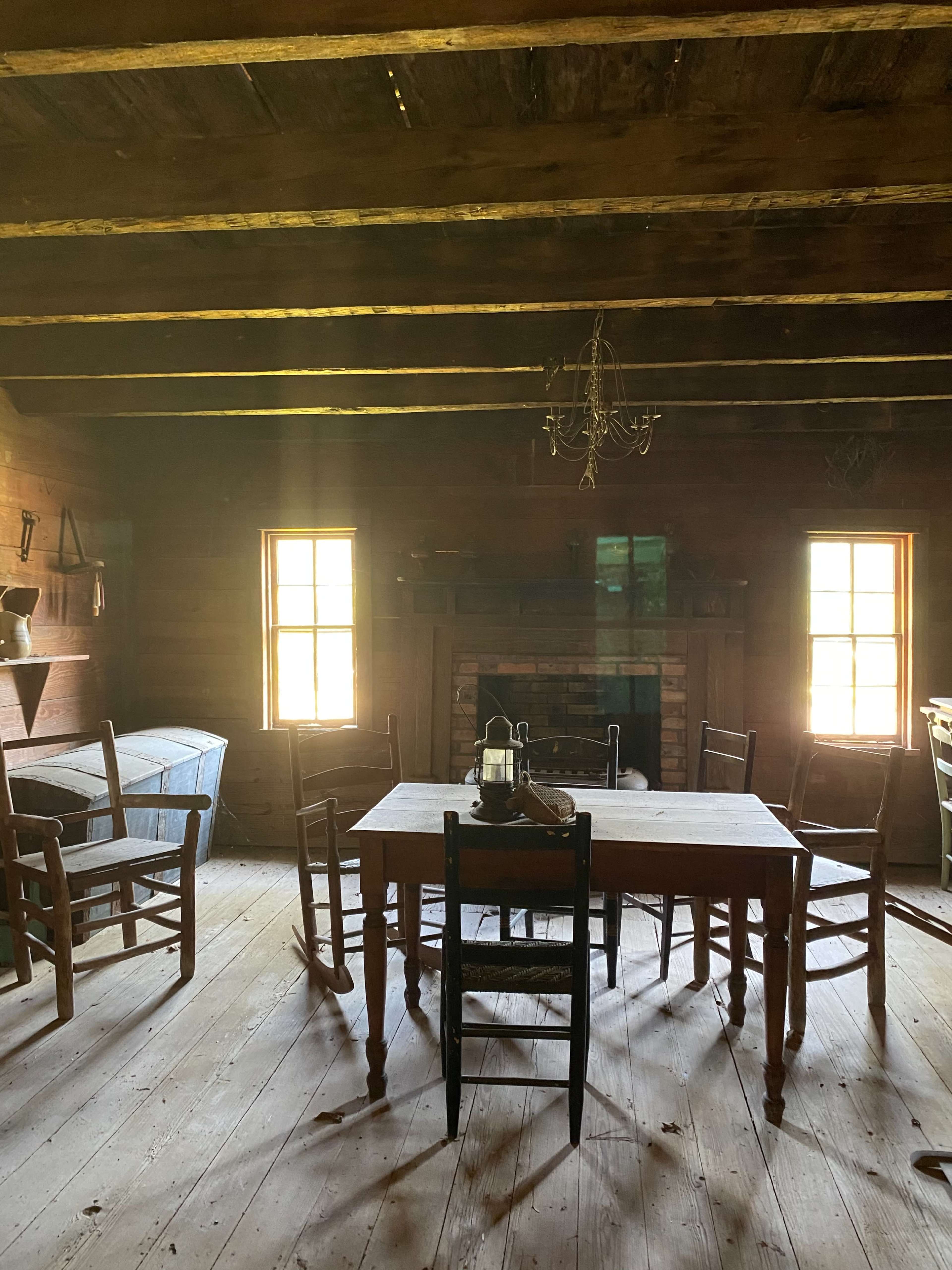 A rustic wooden room with a central table surrounded by several chairs and a fireplace, illuminated by natural light from two windows.