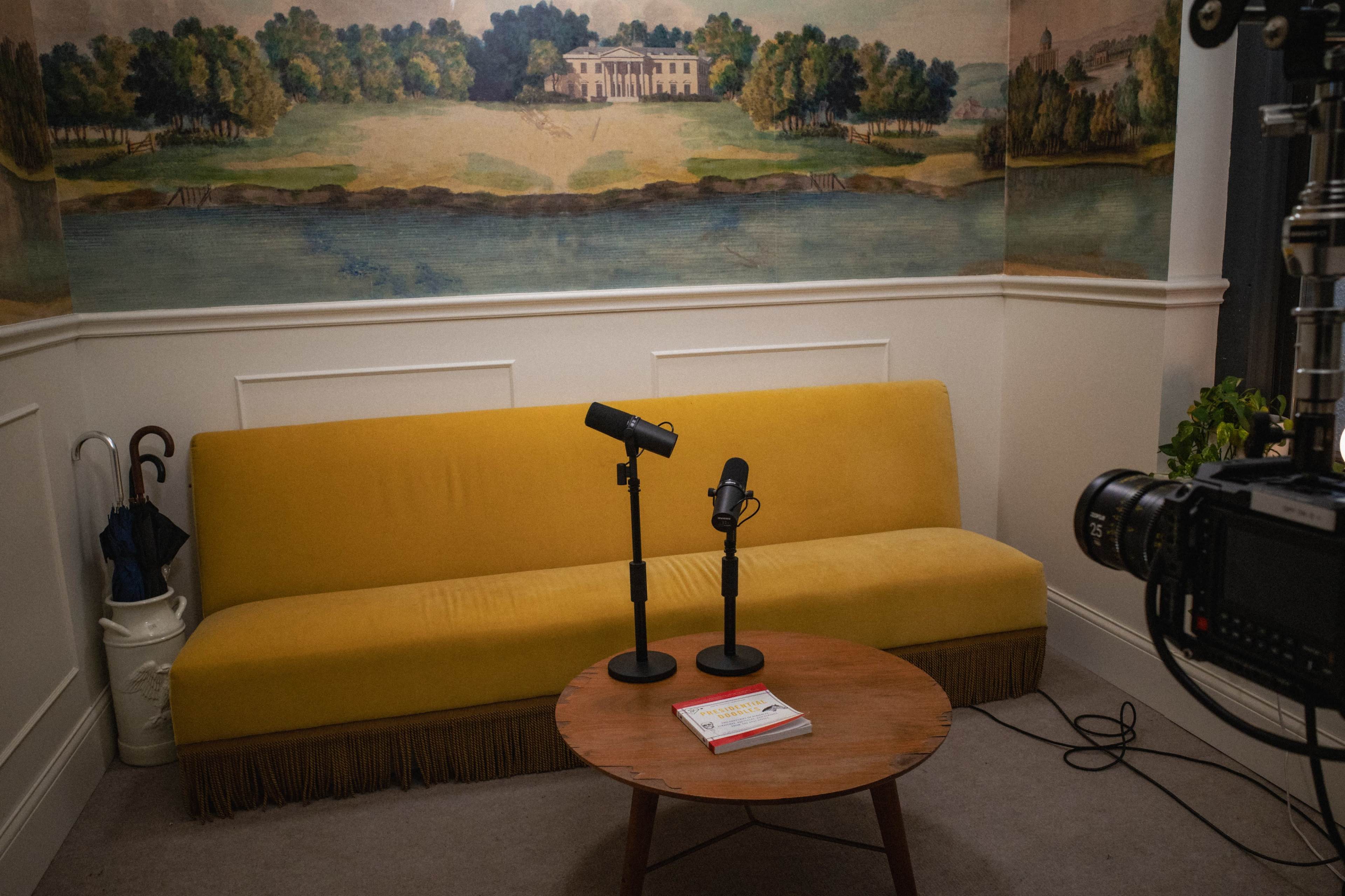 The image shows a yellow sofa and two black microphones positioned on stands in a corner with a round wooden coffee table and a scenic mural depicting a landscape in the background.