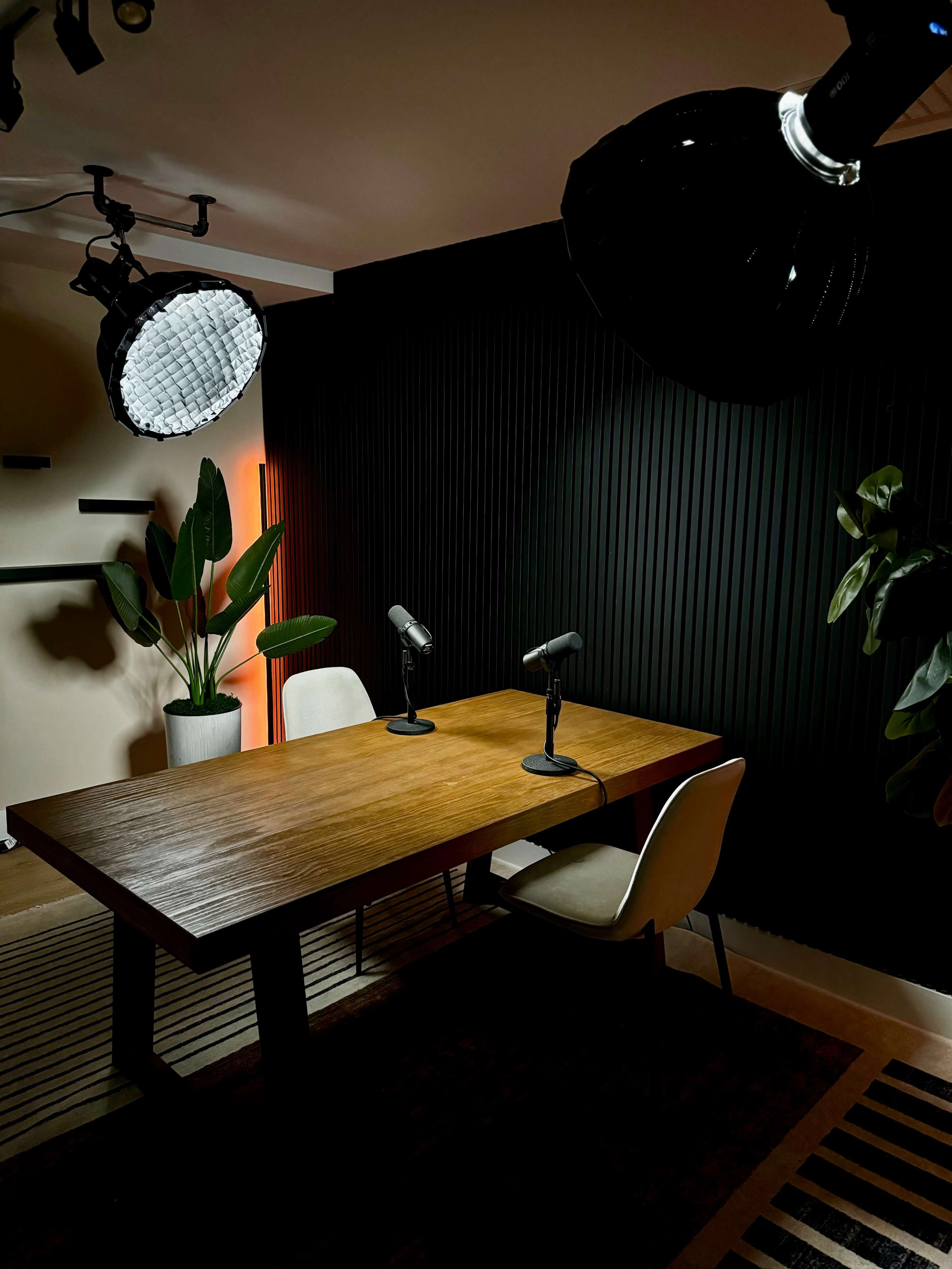 A modern office setting features a wooden table with two microphones, surrounded by chairs and illuminated by studio lights against a dark textured wall.