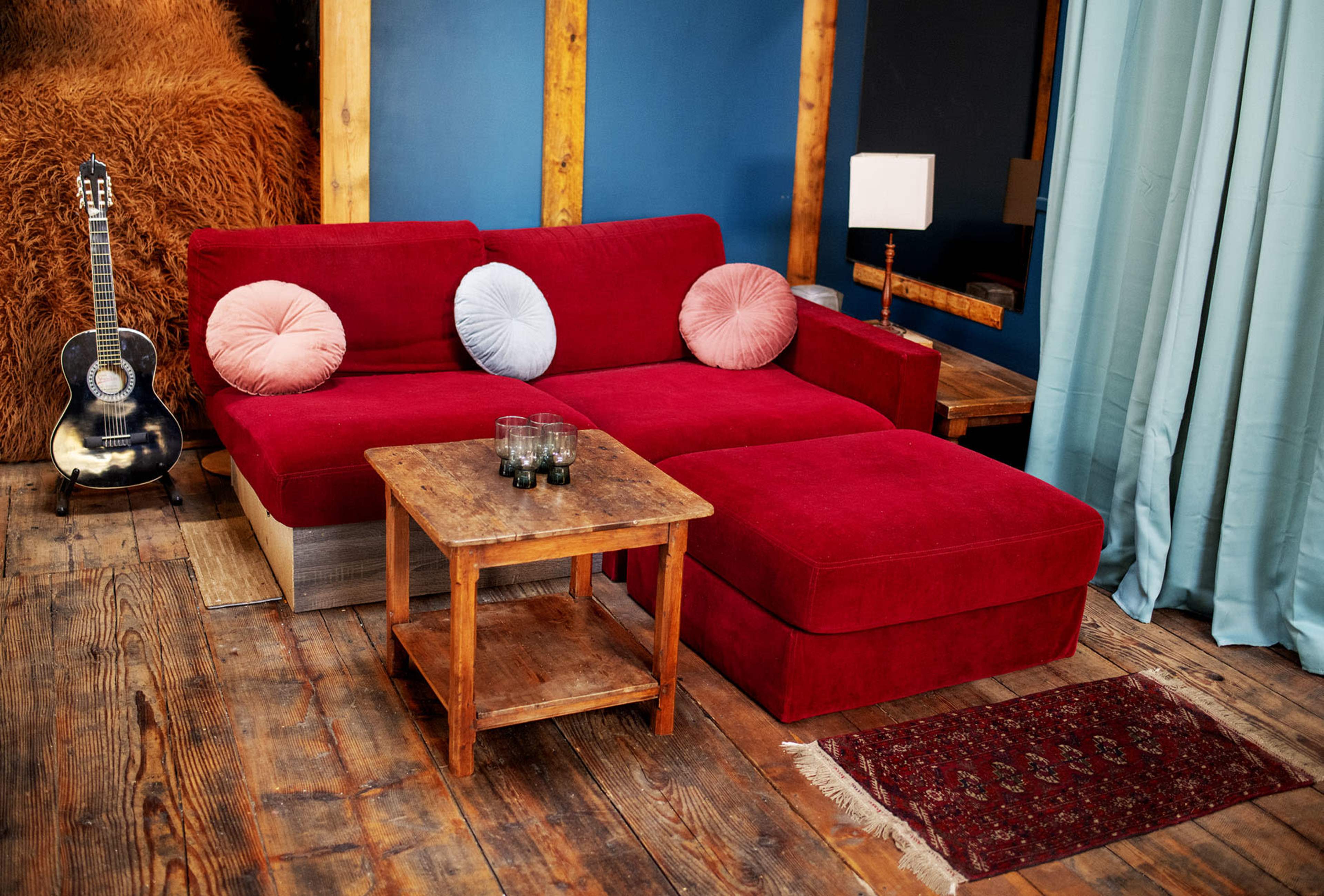 The scene features a red velvet sectional sofa with round cushions, a small wooden coffee table, a guitar in the background, and a curtain separating the space.