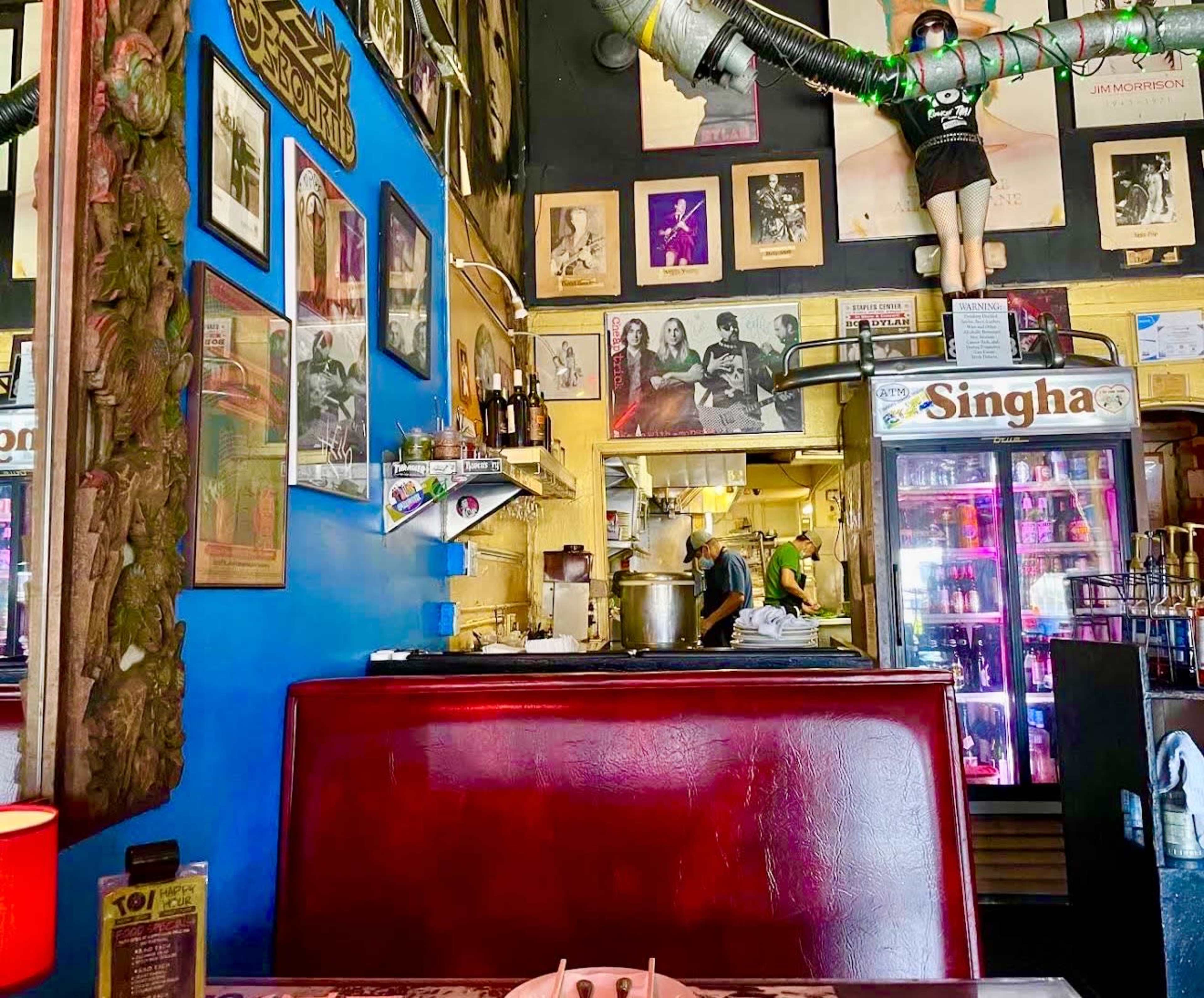 The image shows a vibrant restaurant interior featuring a red booth, eclectic wall art, and a visible kitchen area in the background.