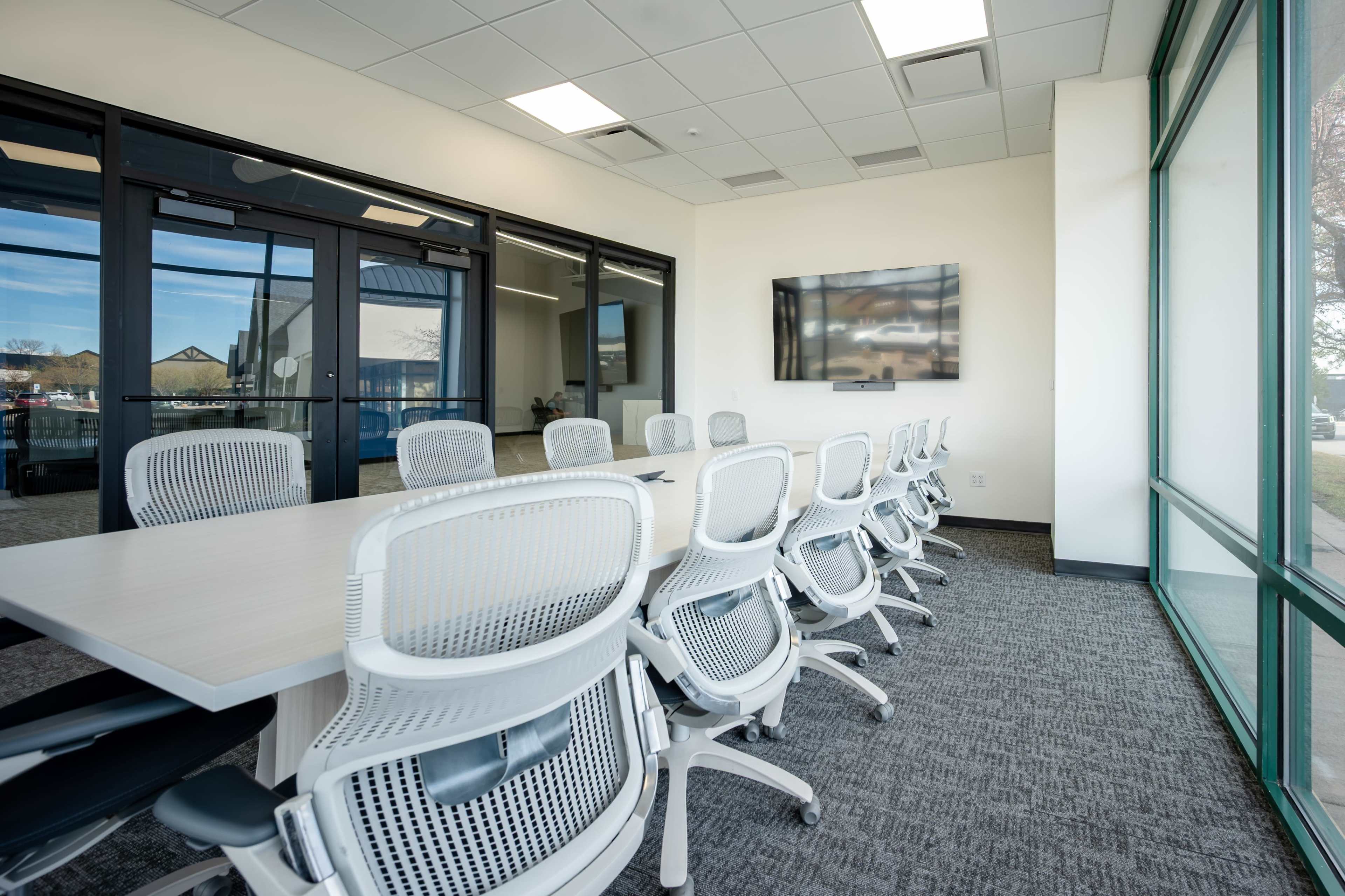 A modern conference room features a long rectangular table surrounded by white ergonomic chairs and a wall-mounted television.
