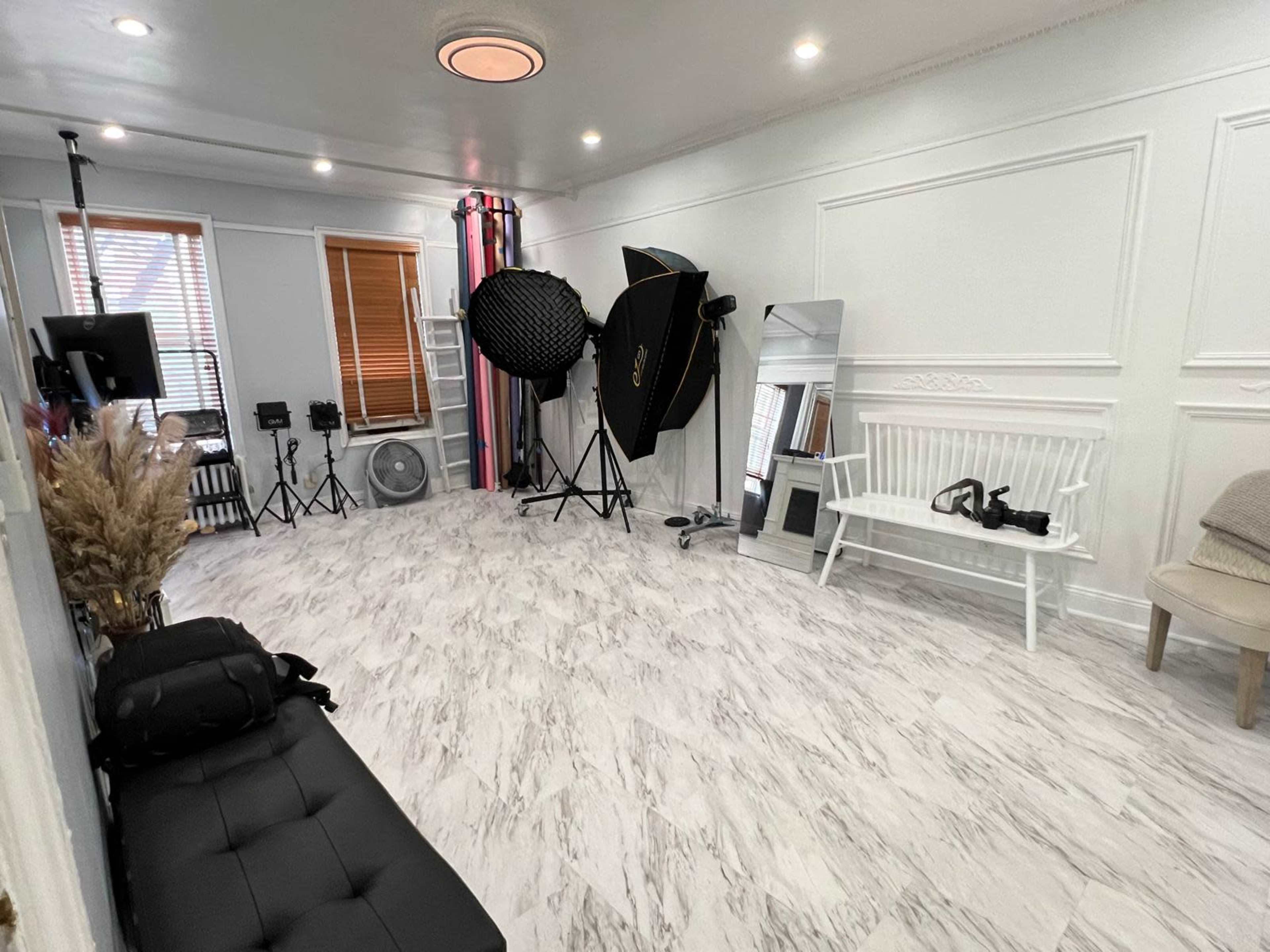 The image shows a photography studio with a light-colored marble floor, studio equipment such as softboxes and a reflector, and furniture including a bench and a chair.