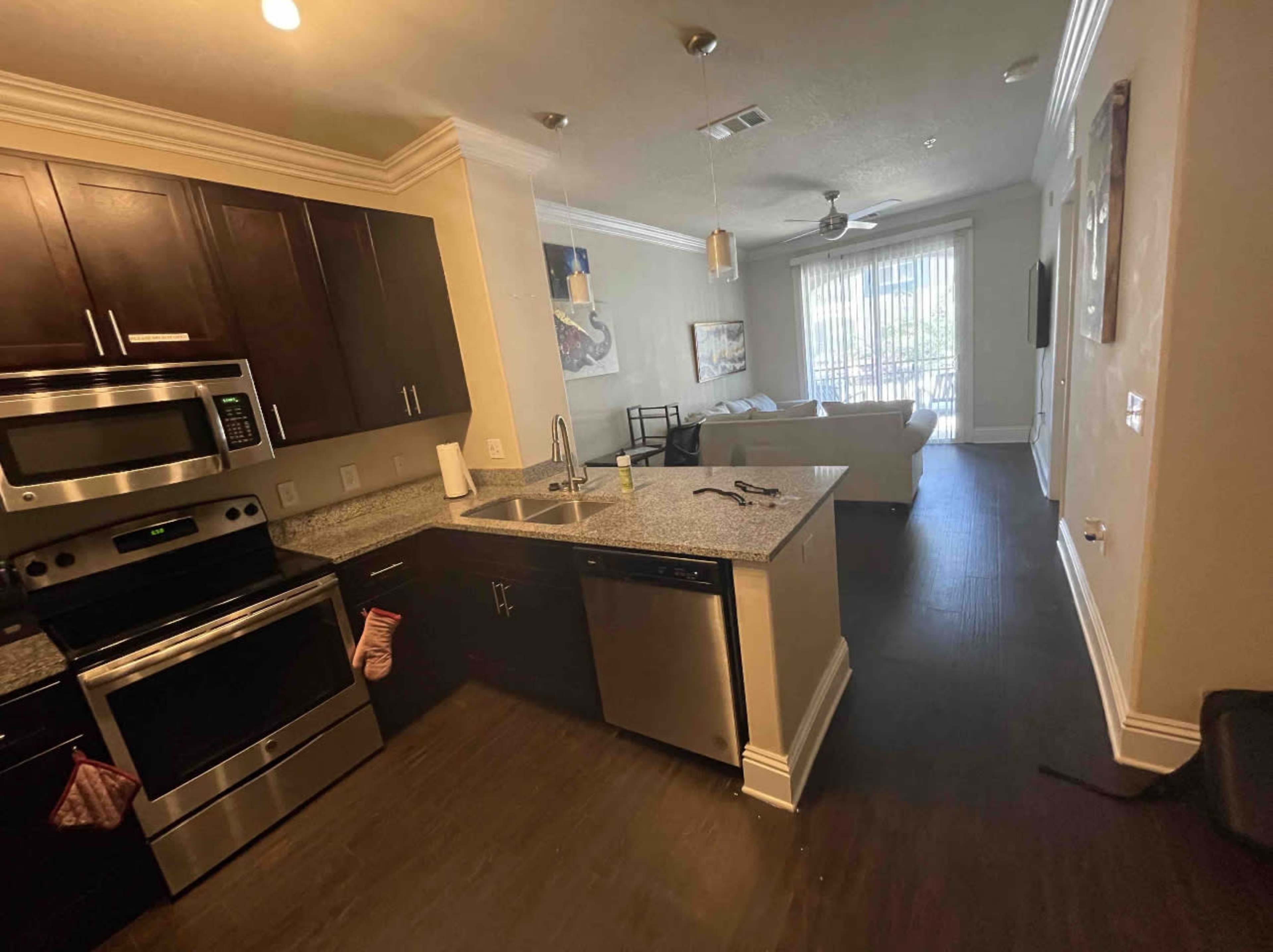 The image shows a modern kitchen with dark cabinets and stainless steel appliances, adjacent to a living area with a sofa and large windows.
