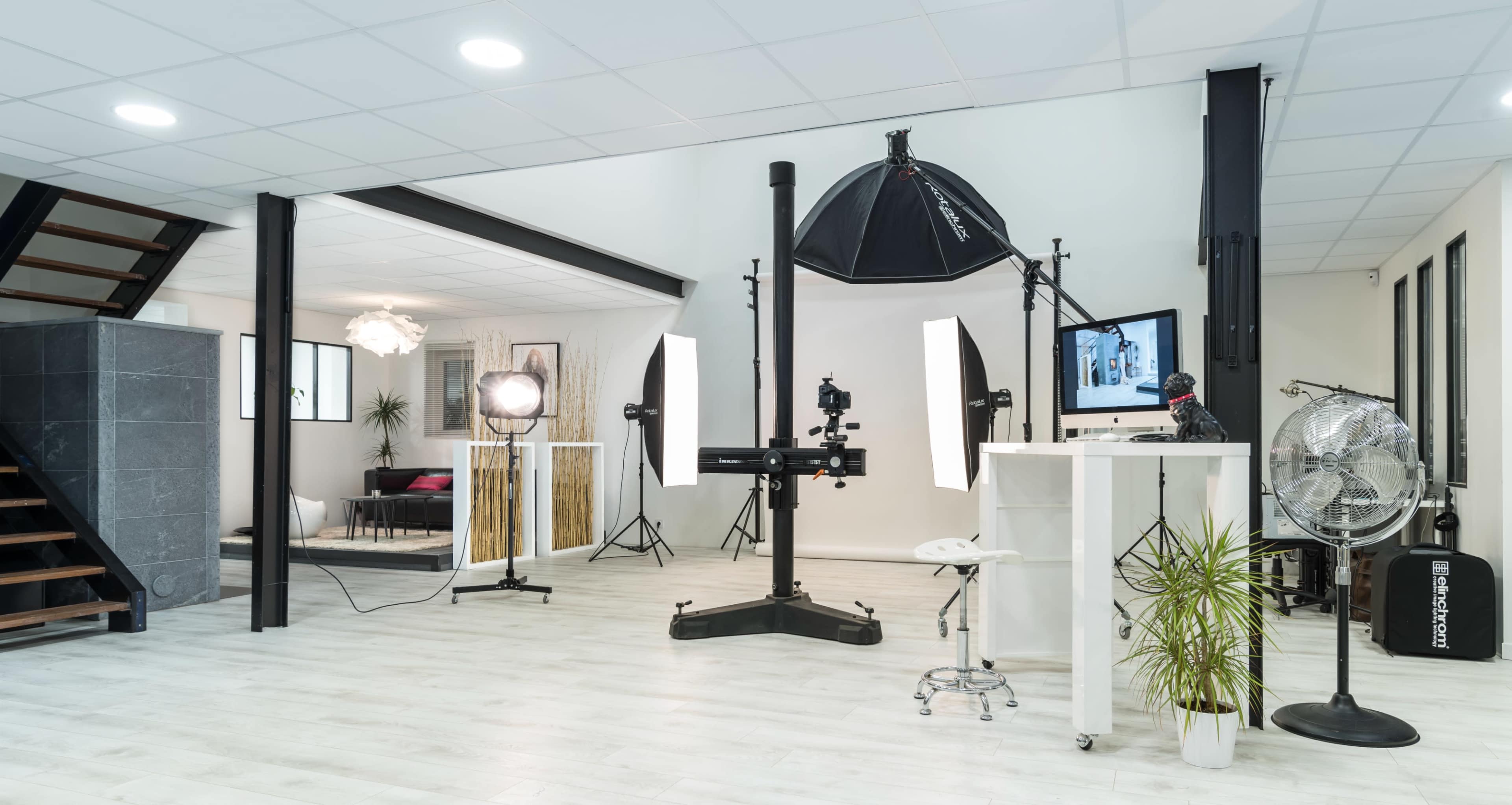 The image shows a modern photography studio with a variety of lighting equipment, a computer setup, and decorative plants.