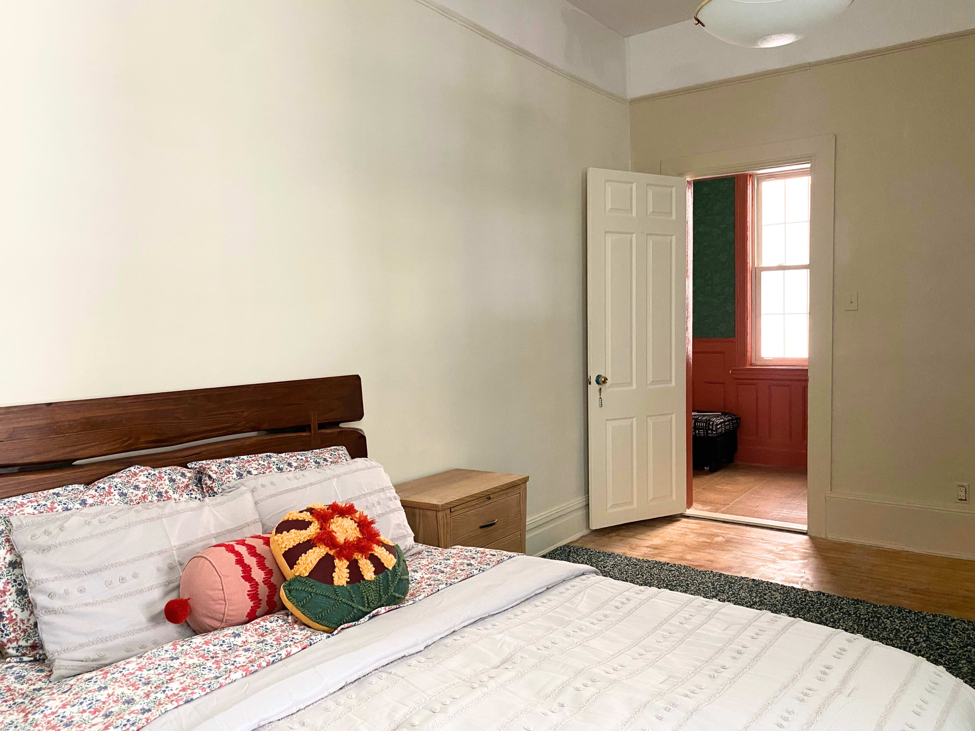 A neatly made bed with decorative pillows is positioned against a wall, and an open door leads to another room with visible woodwork and a window.