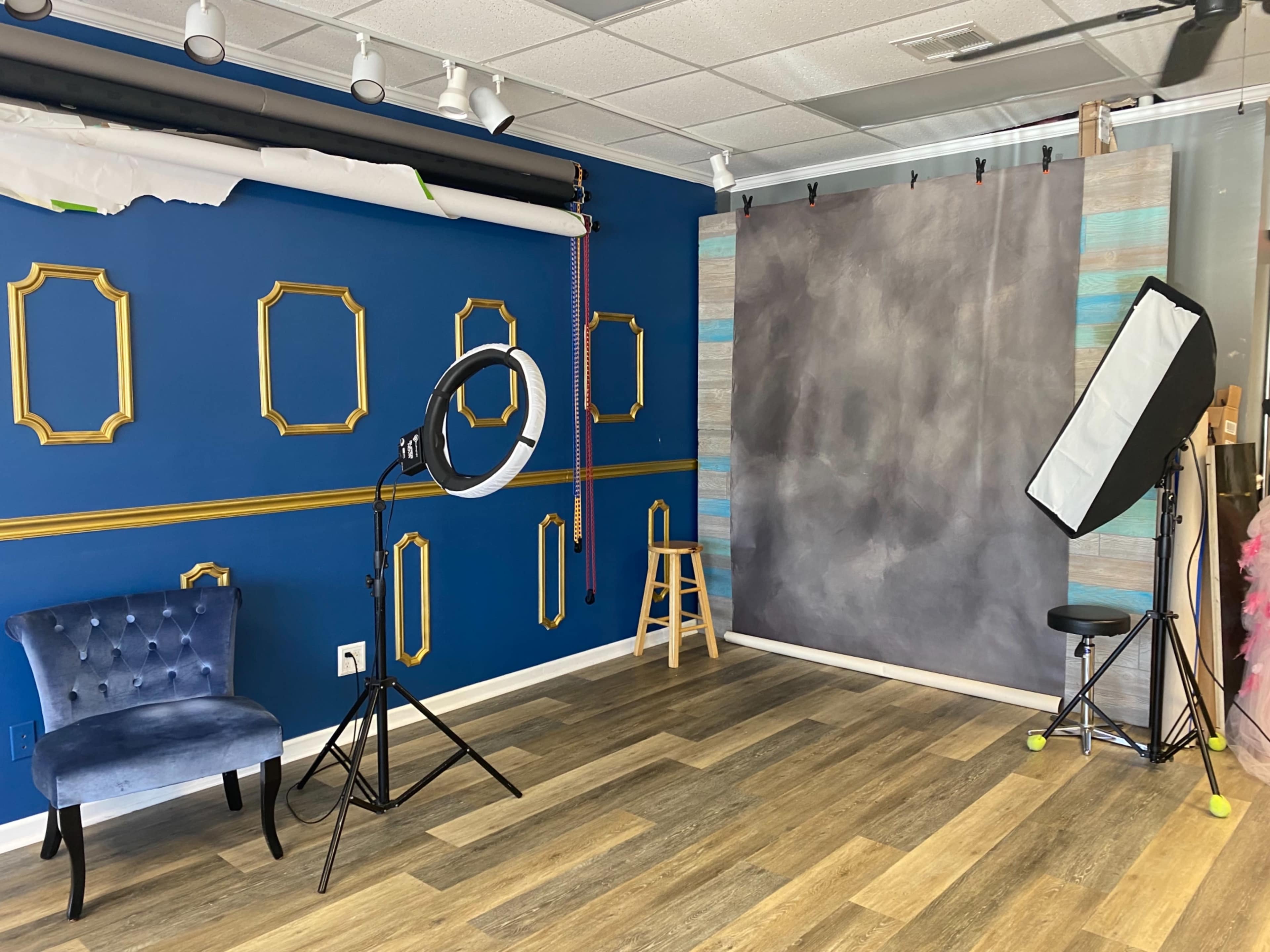 A photography studio with a blue wall featuring decorative frames, a gray backdrop, lighting equipment, and a blue velvet chair.