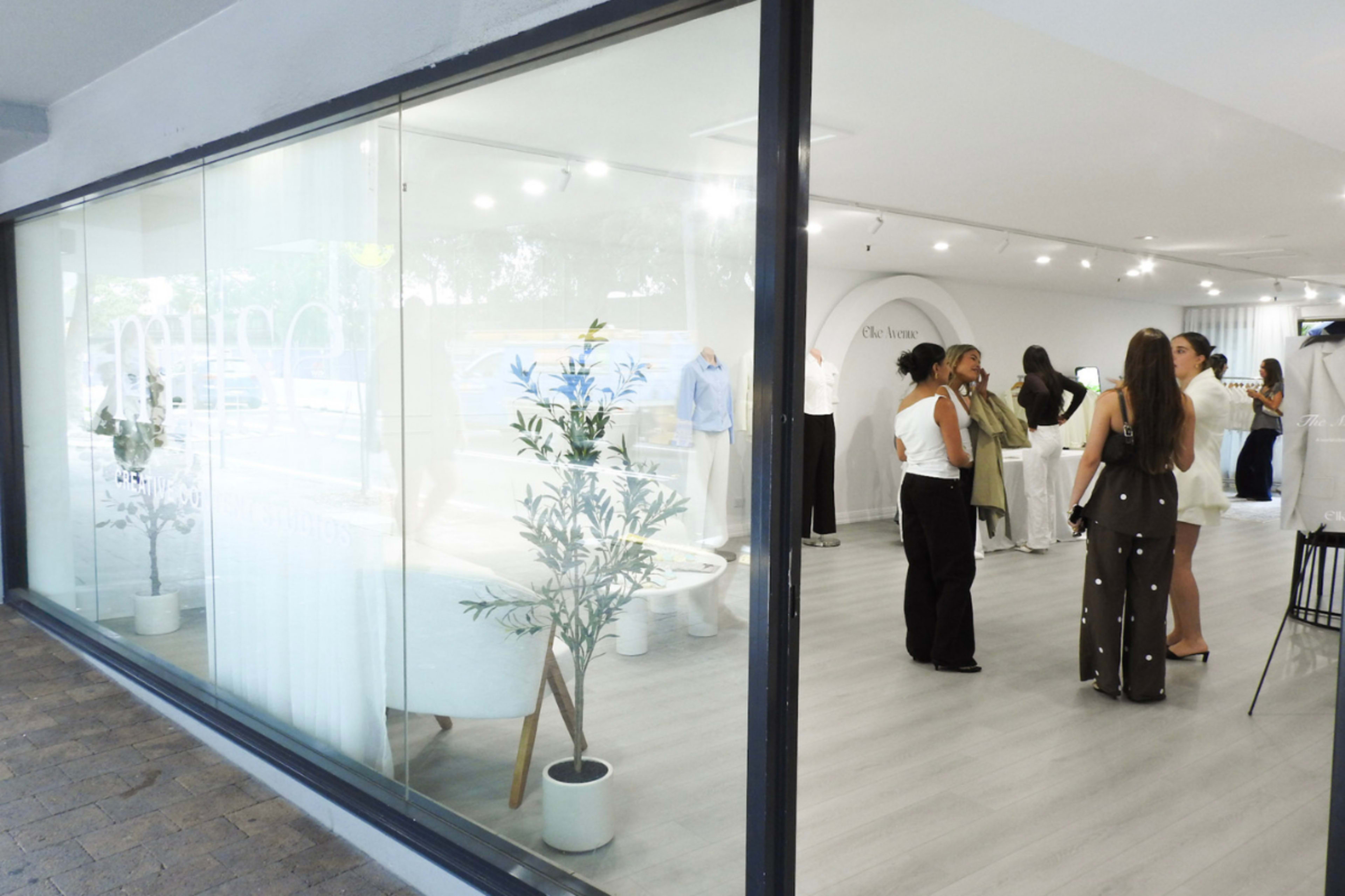 The image shows a modern boutique with large windows, featuring several people engaged in conversation and browsing clothing items inside.