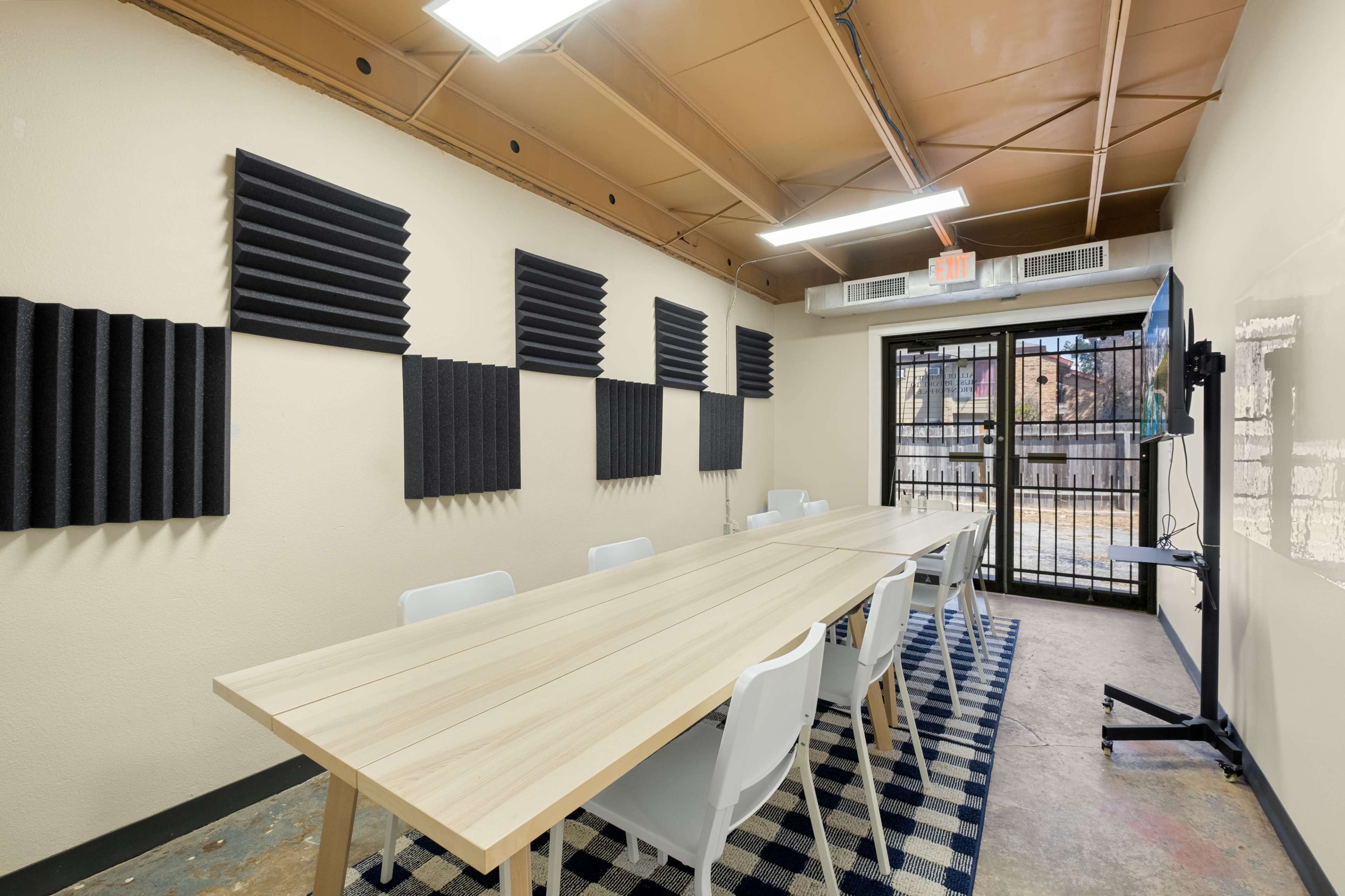 A long wooden table surrounded by white chairs is located in a meeting room with sound-absorbing panels on the walls and a large window with a metal grate.