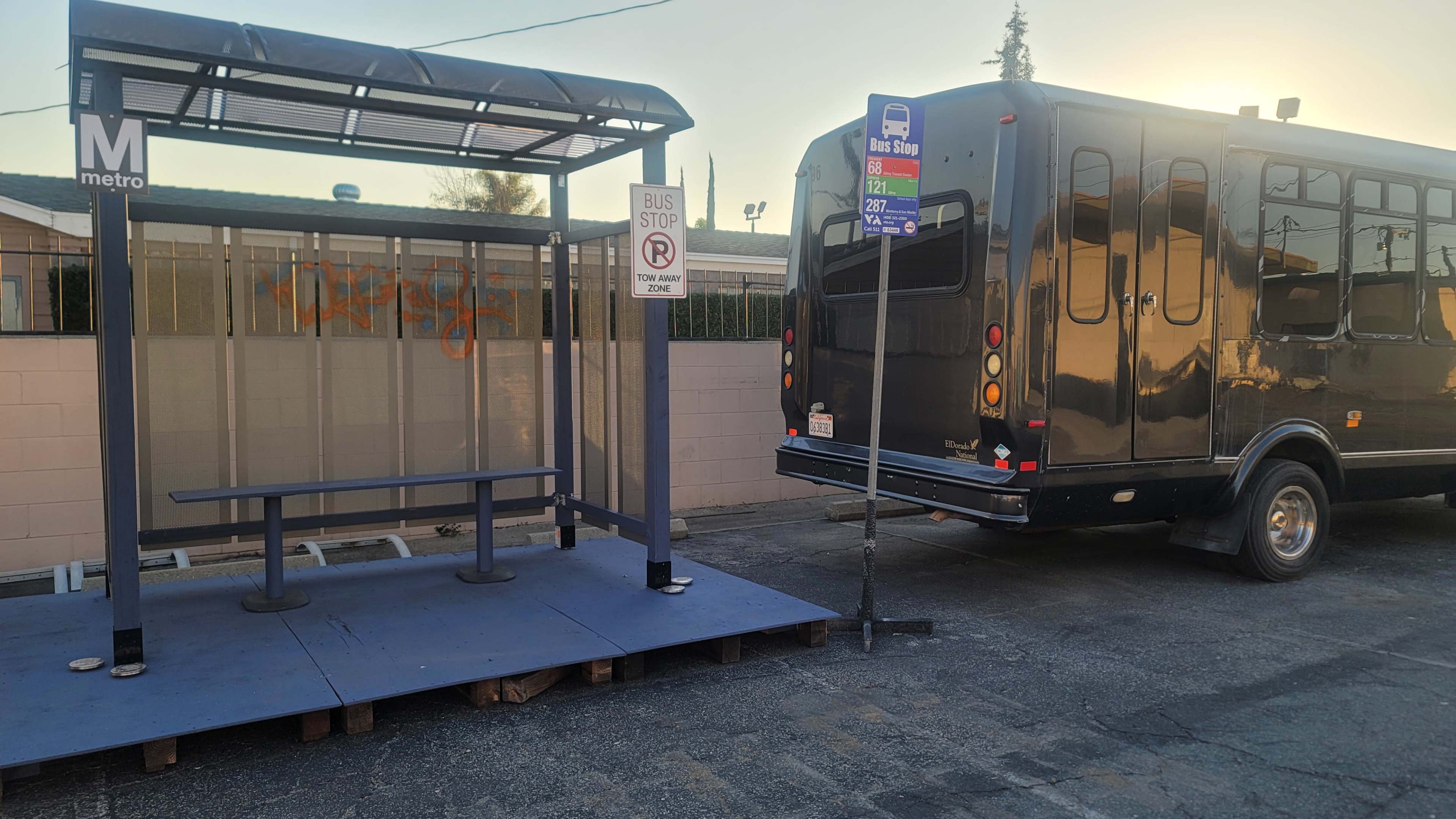 A bus stop structure with a bench is positioned beside a parked transit bus on a city street.
