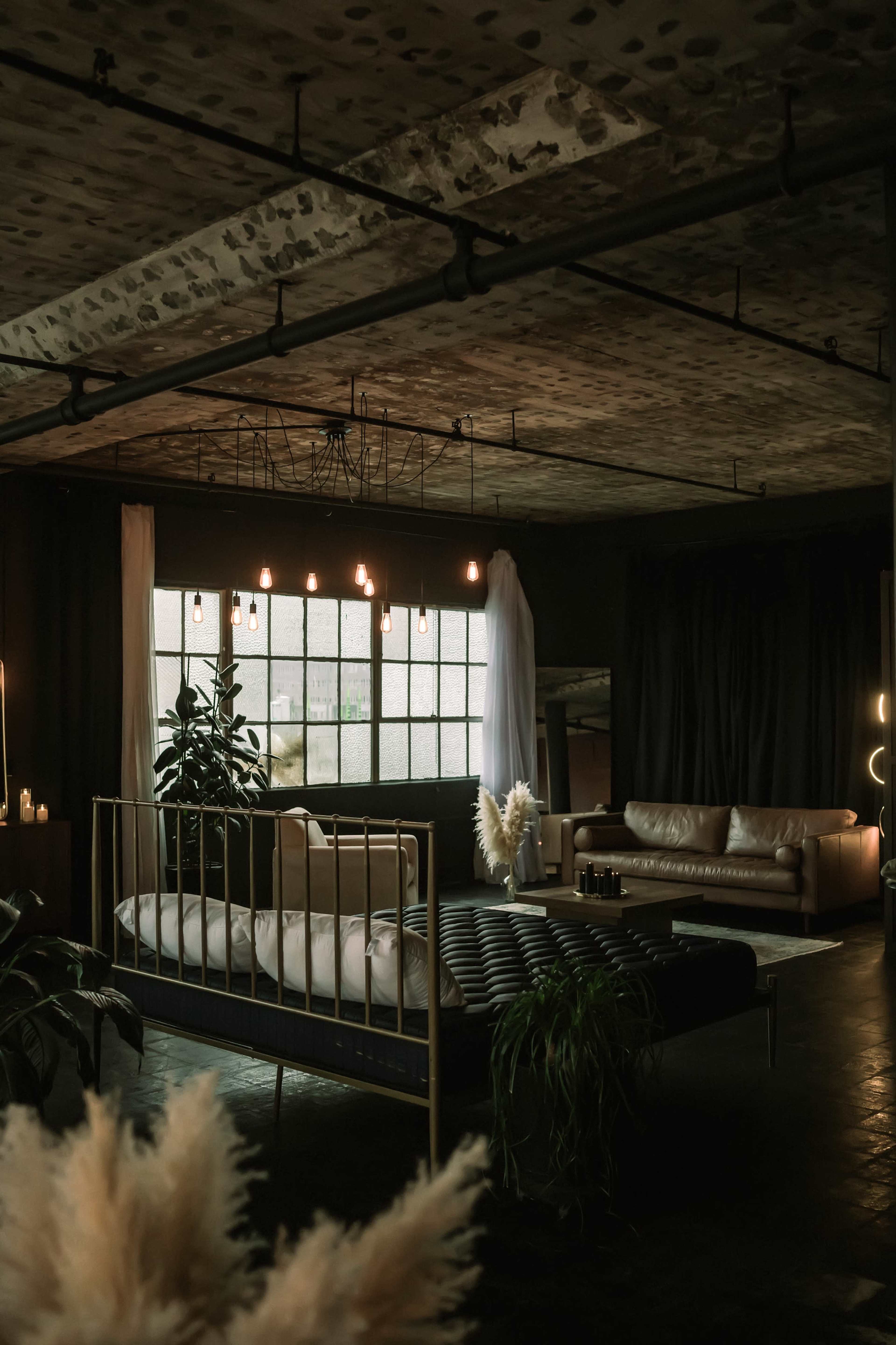 The image shows a modern interior space featuring a bed with a gold frame, a leather sofa, large windows with curtains, and various plants.