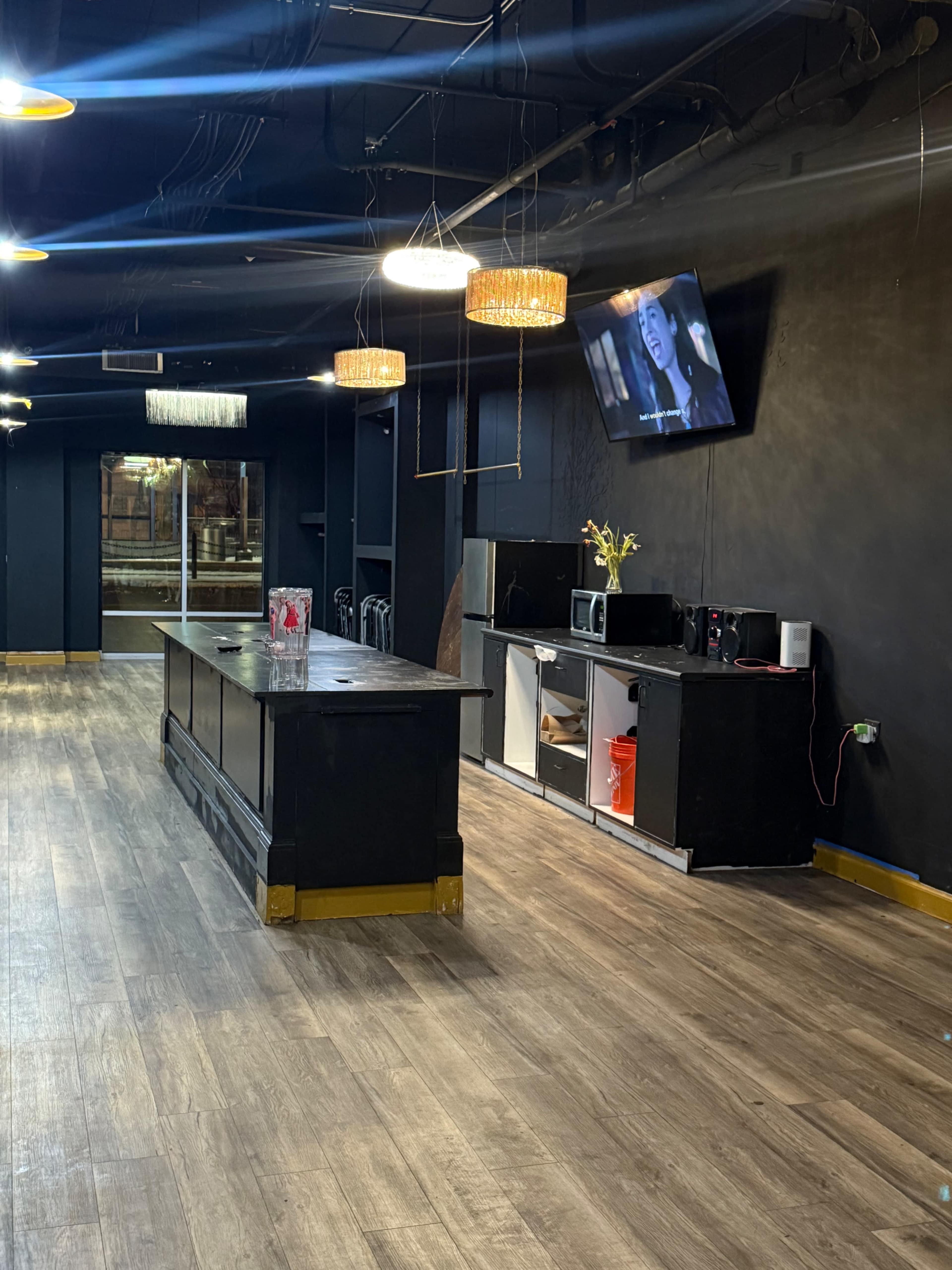 The image shows a modern, dimly lit space featuring a long black bar counter, several appliances, and a television mounted on the wall.