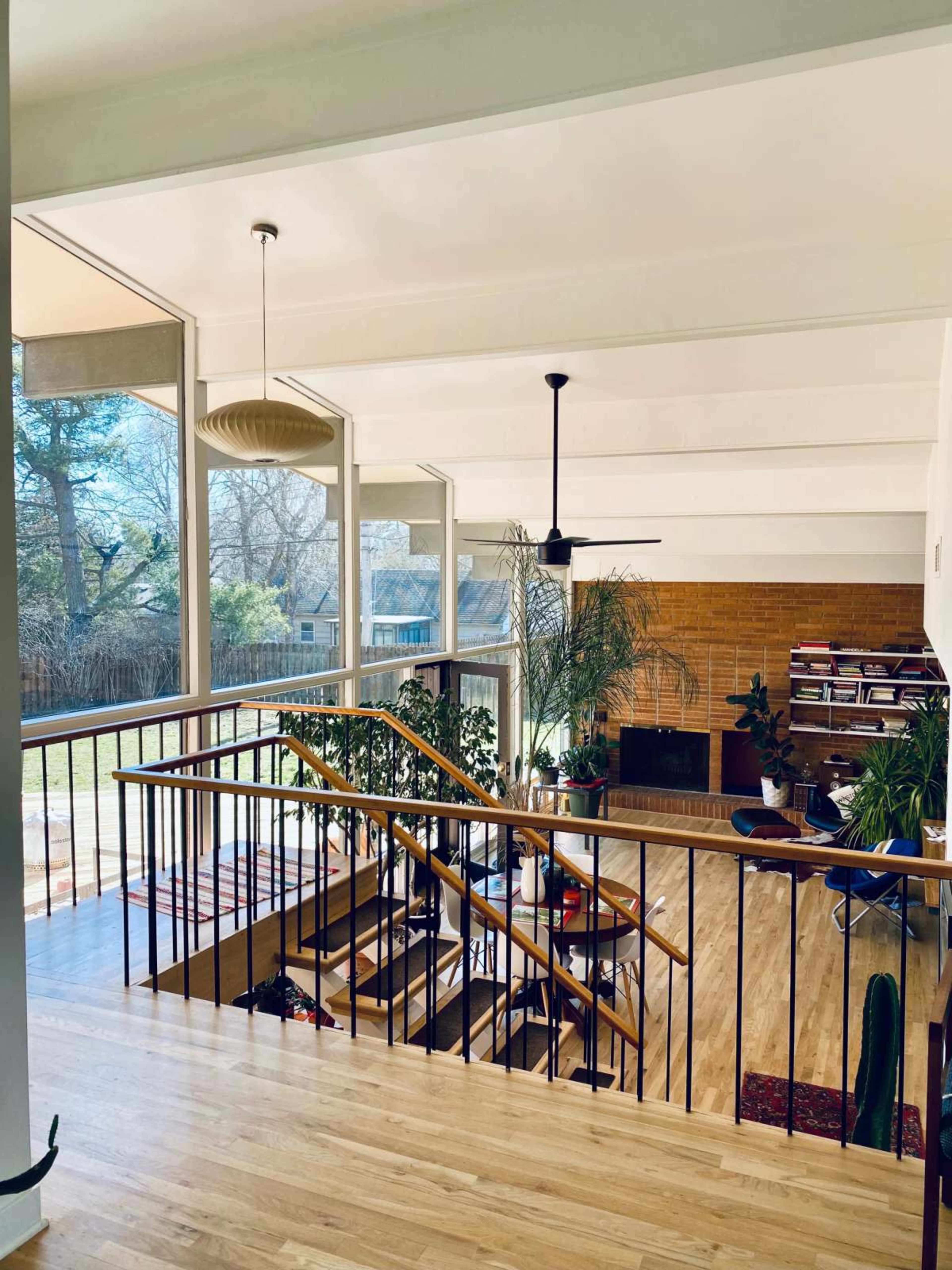 The image shows a spacious living area with large windows, a wooden staircase, and a brick fireplace surrounded by plants and bookshelves.