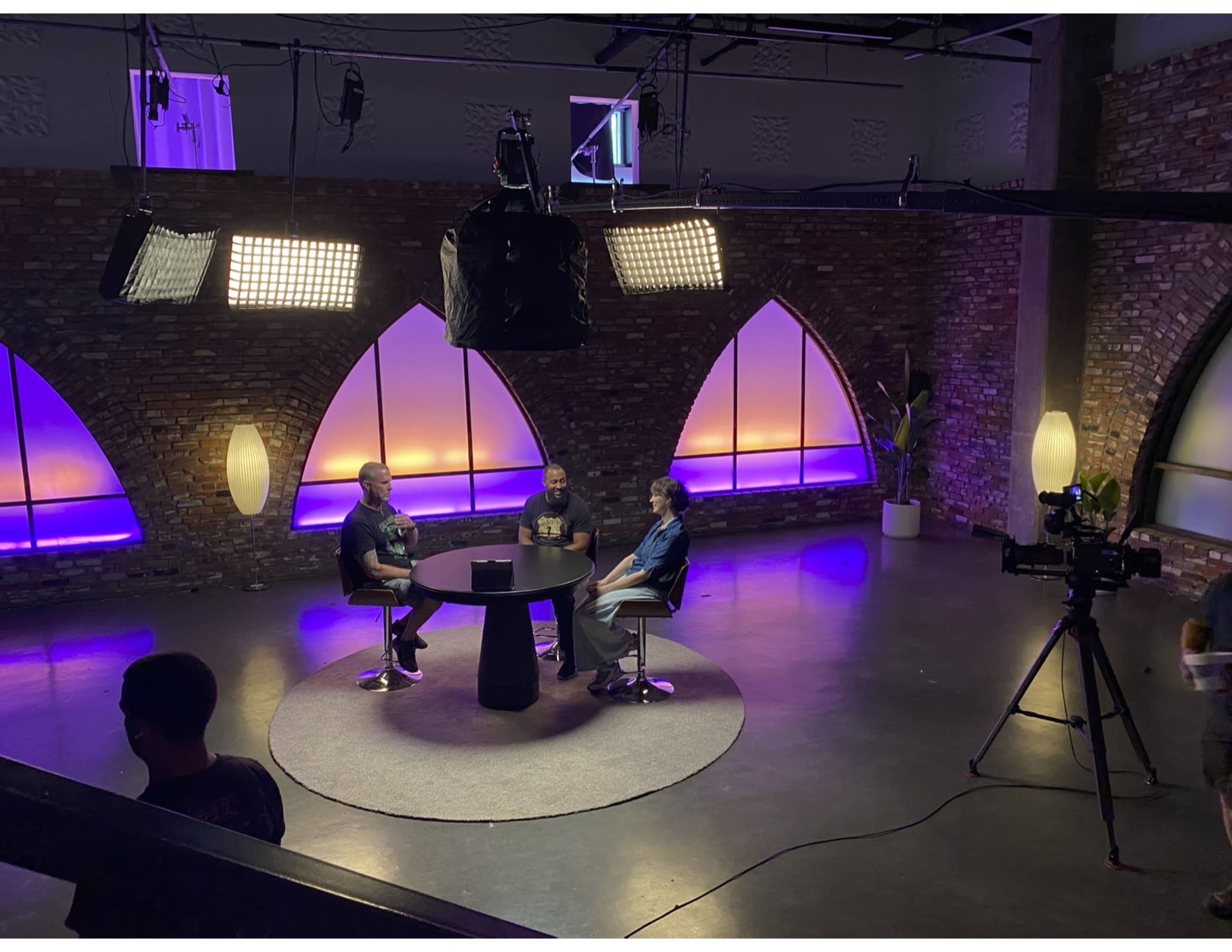 Three people sit at a round table in a studio with colorful lighting and a camera set up nearby.