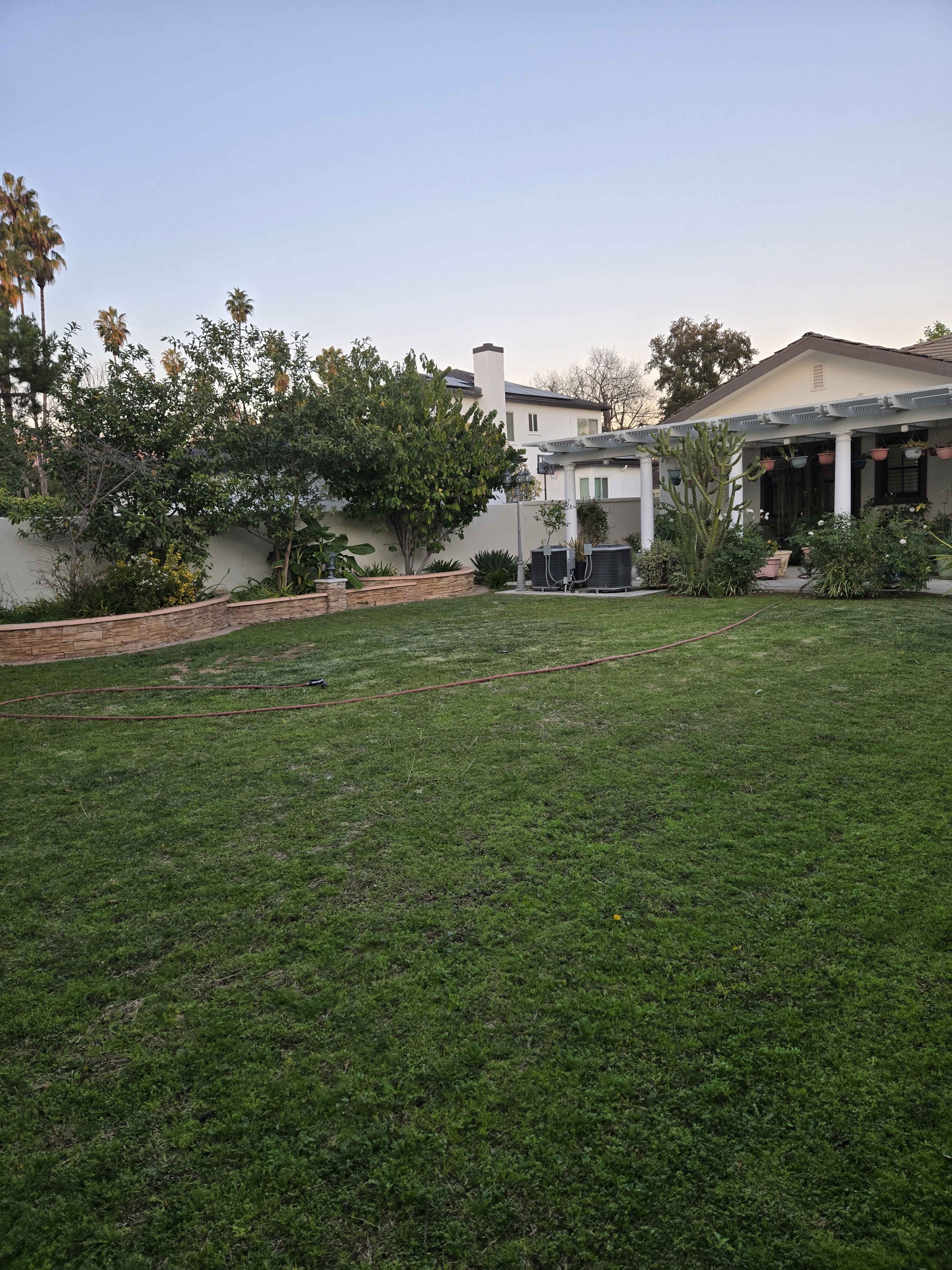 The image shows a spacious green lawn with garden beds, a patio area, and trees in a residential backyard.
