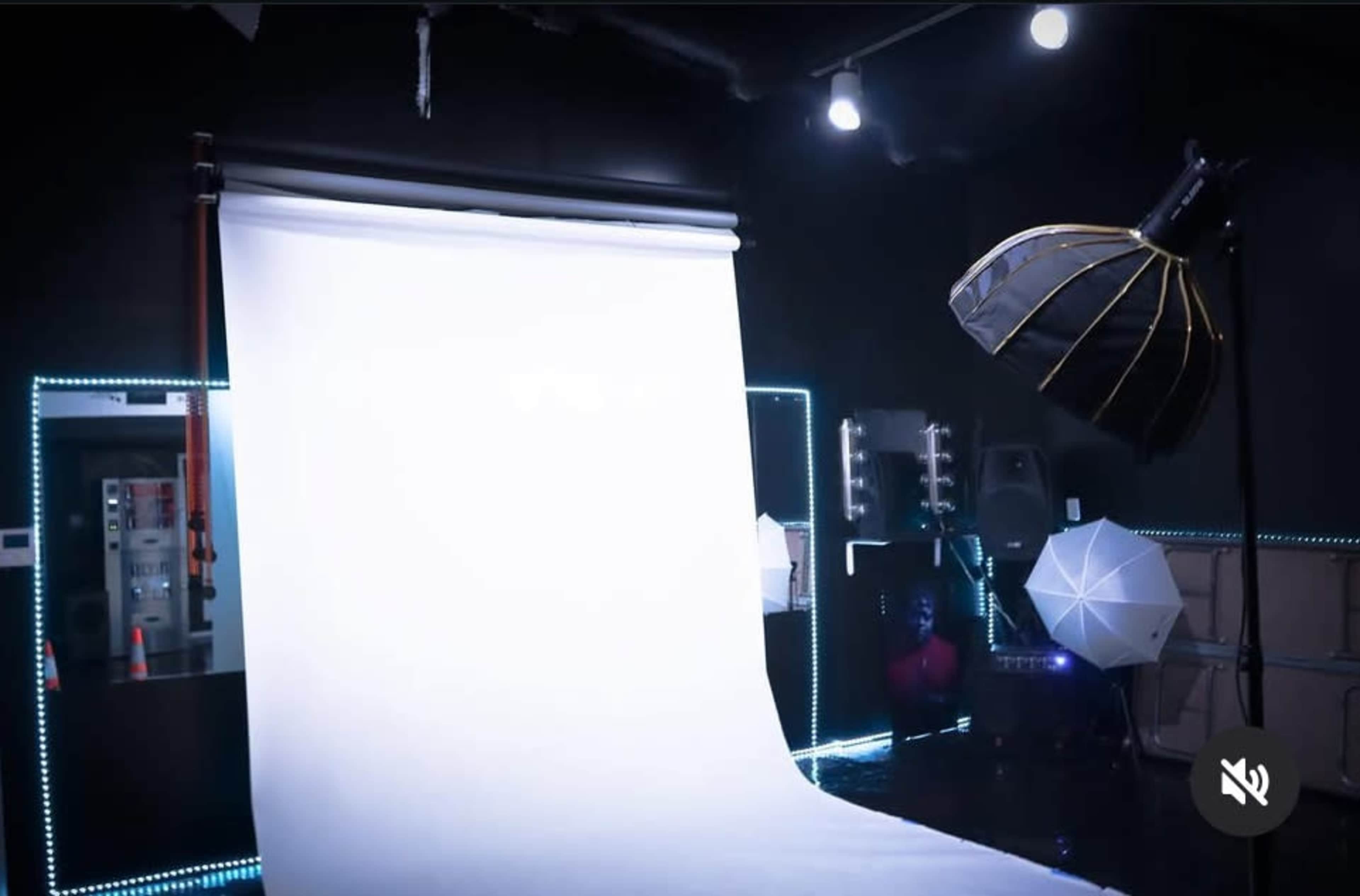 The image shows a photography studio with a white backdrop, lighting equipment, and reflectors arranged for a photoshoot.
