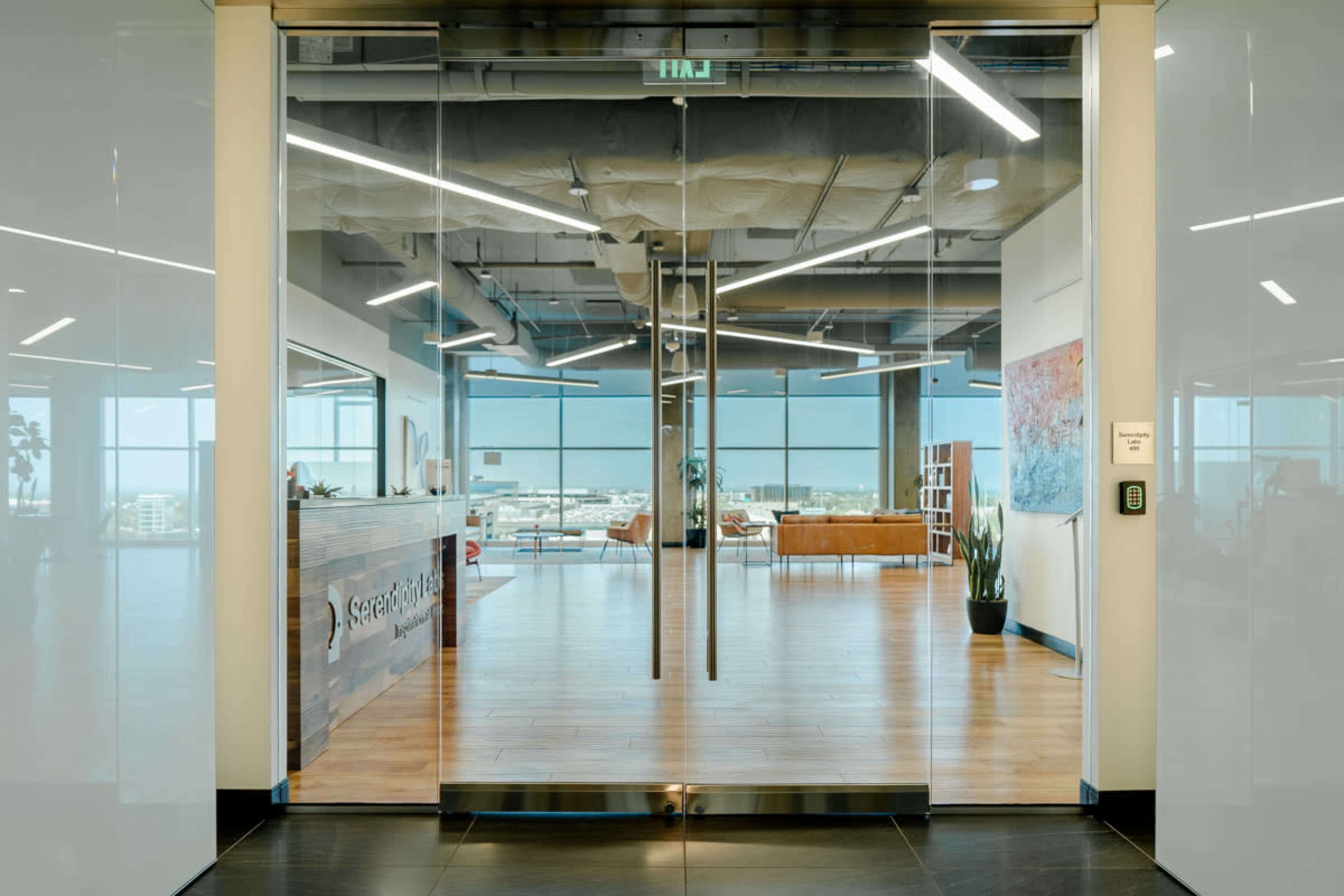 The image shows a modern office lobby with glass doors leading to an open space featuring a reception desk, seating area, and large windows overlooking a cityscape.
