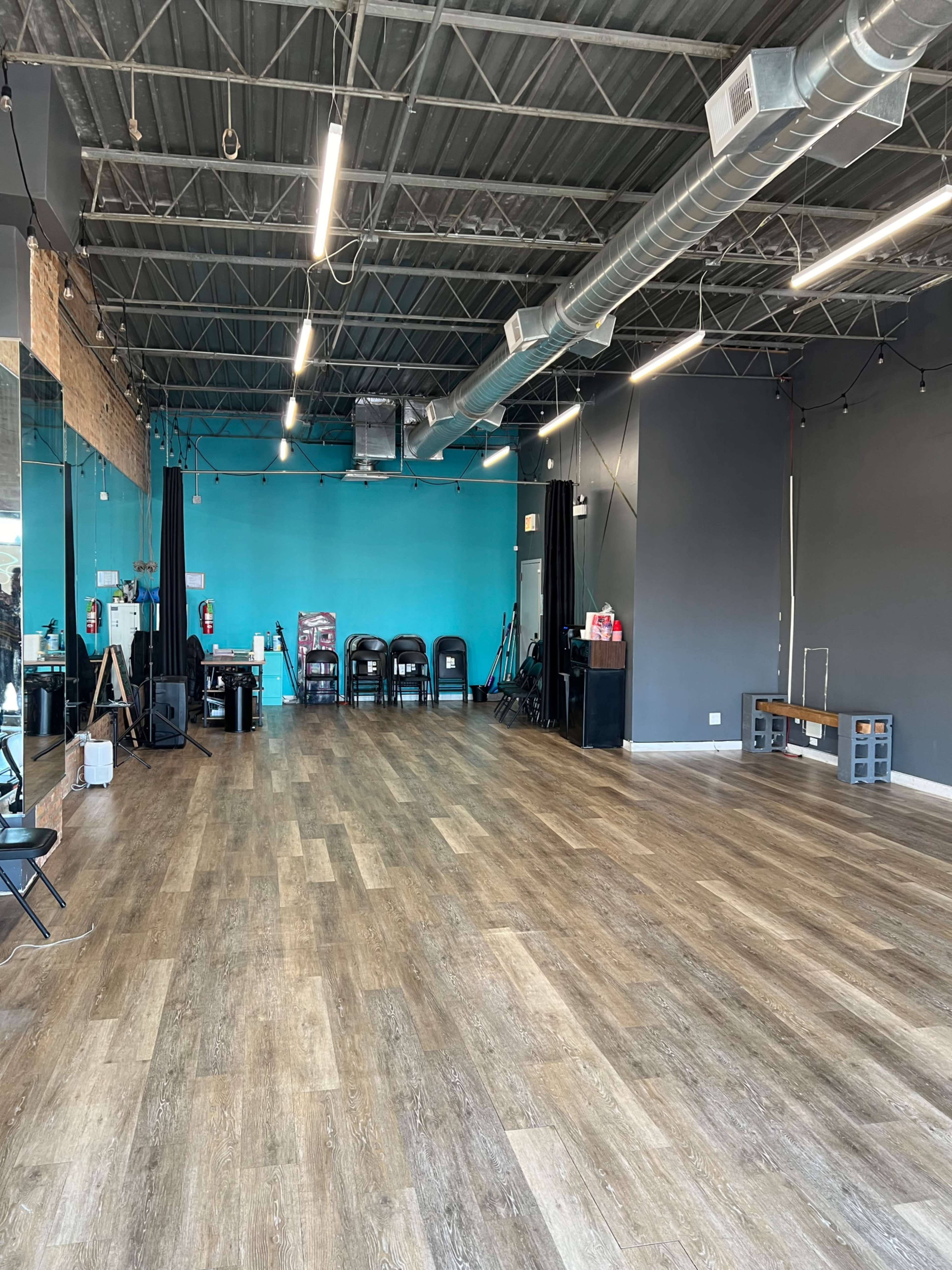 A spacious studio with wooden flooring, teal and gray walls, and various chairs and equipment along the sides.