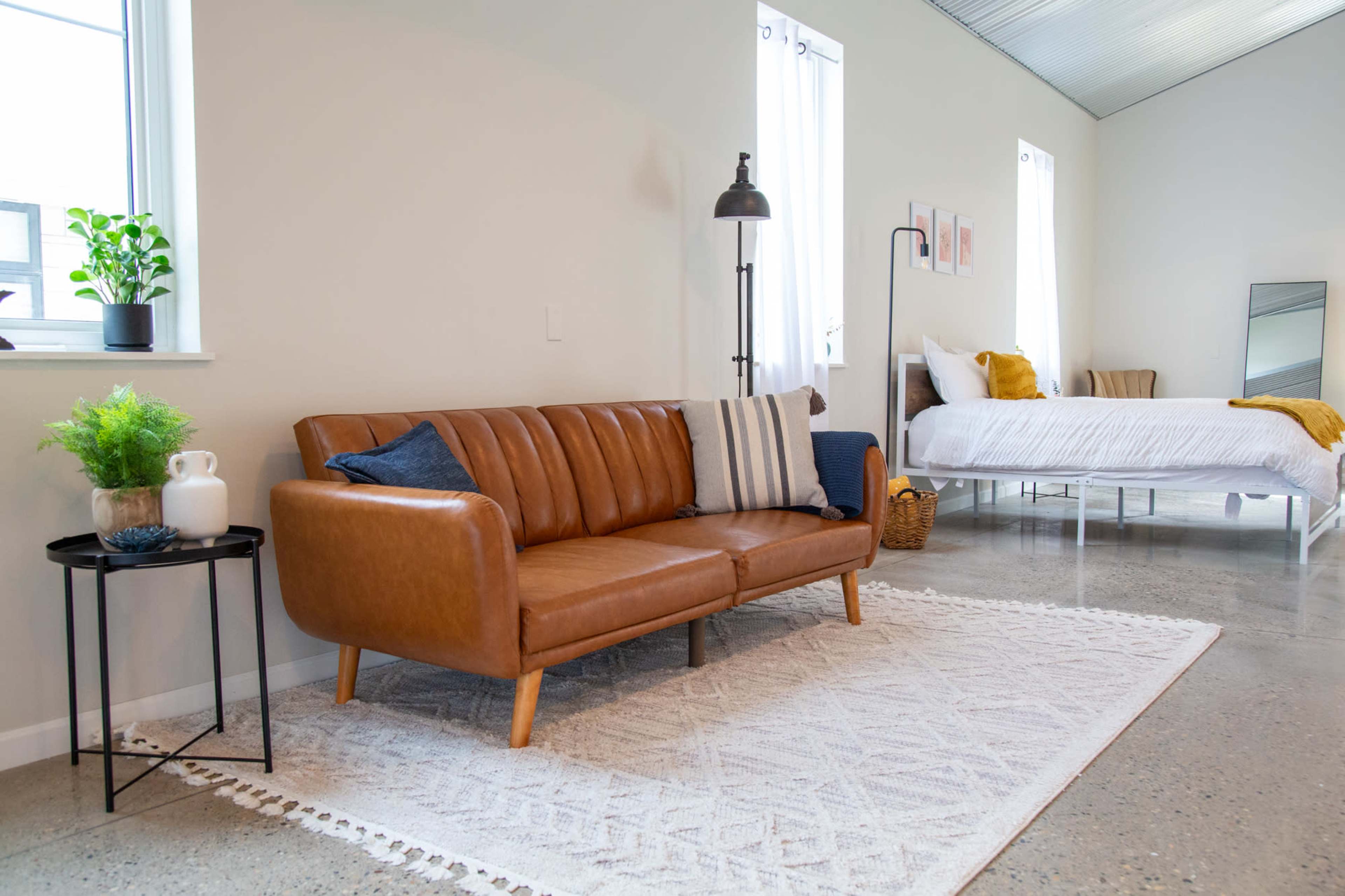 A modern living space featuring a brown leather sofa, a black side table with a plant, and a bedroom area with a white bed and a large mirror.