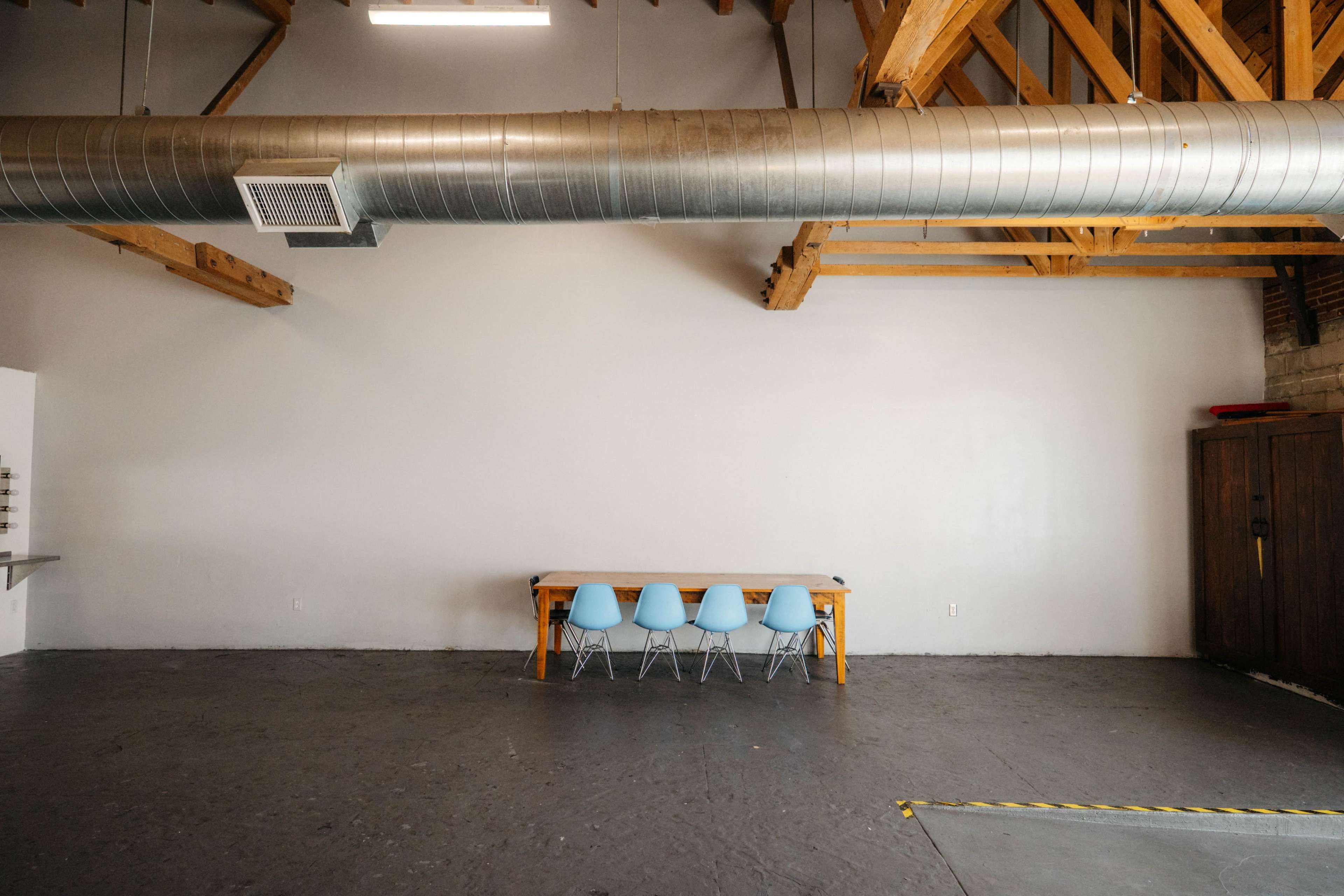 The image shows a spacious room with a long wooden table and four blue chairs against a plain wall, featuring exposed wooden beams and a large metal duct overhead.