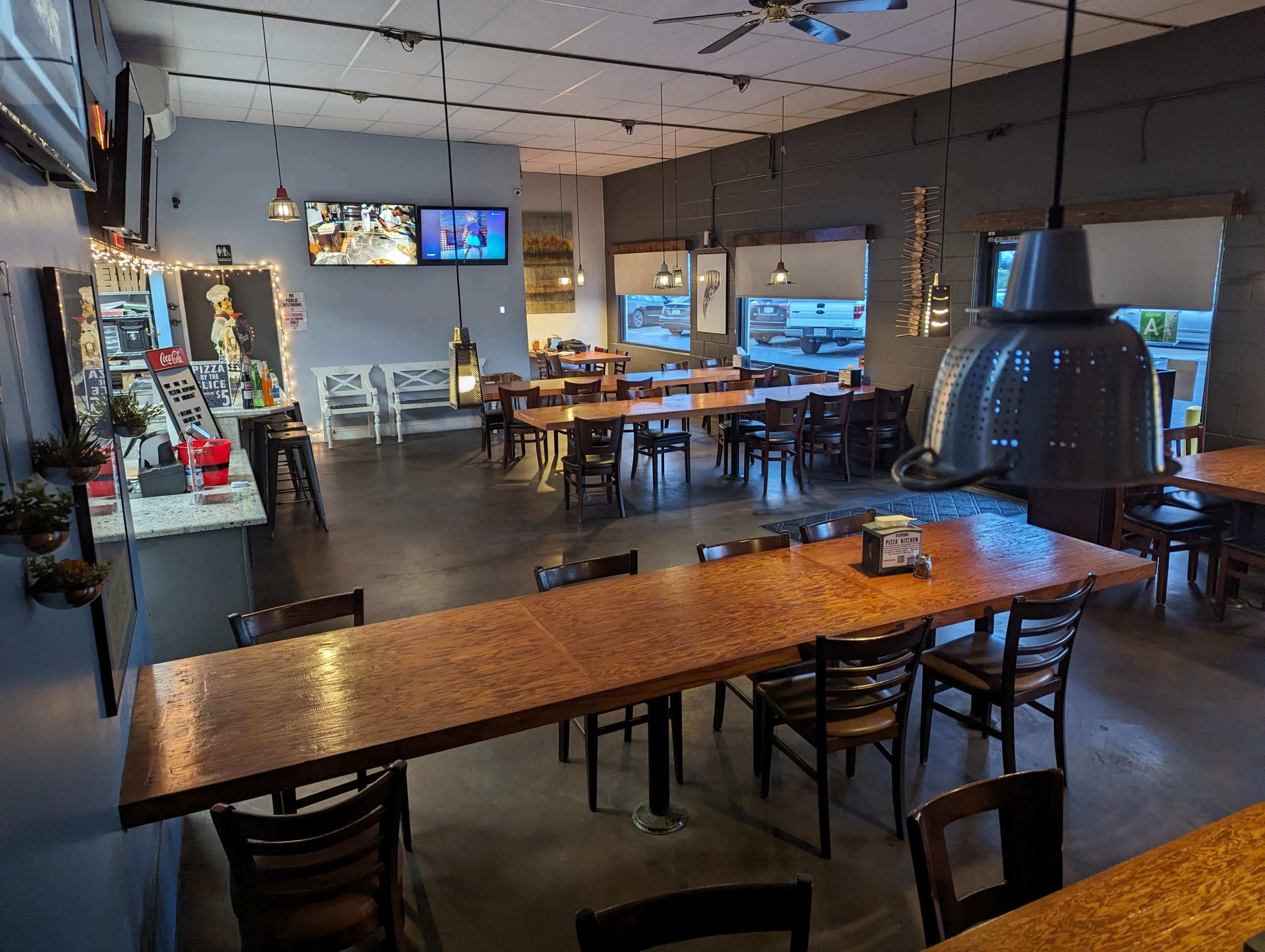 An indoor dining area of a restaurant with wooden tables, chairs, and a wall-mounted TV, featuring a counter area and large windows.