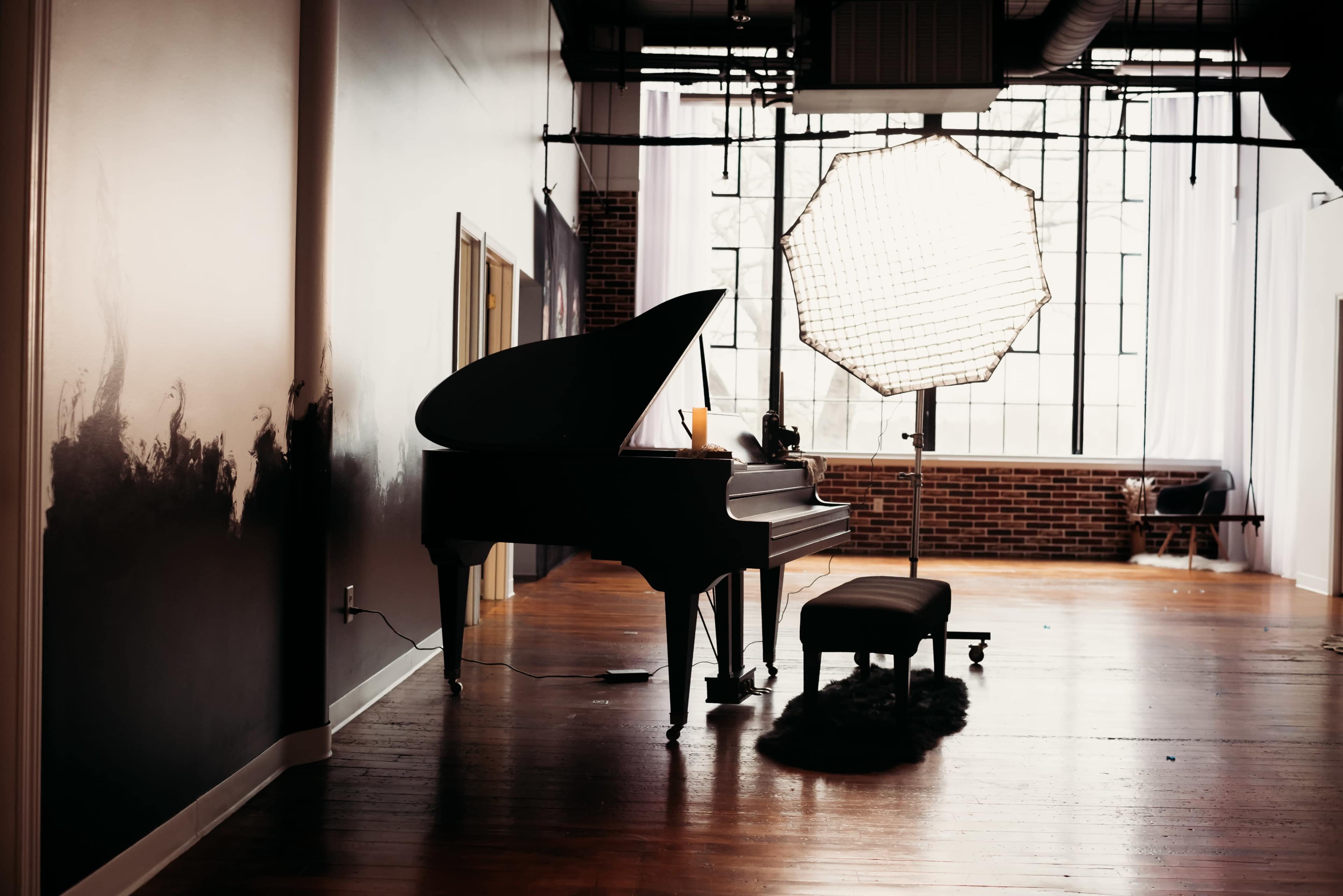 A grand piano is positioned next to a large window, with a softbox light situated nearby in a spacious room.