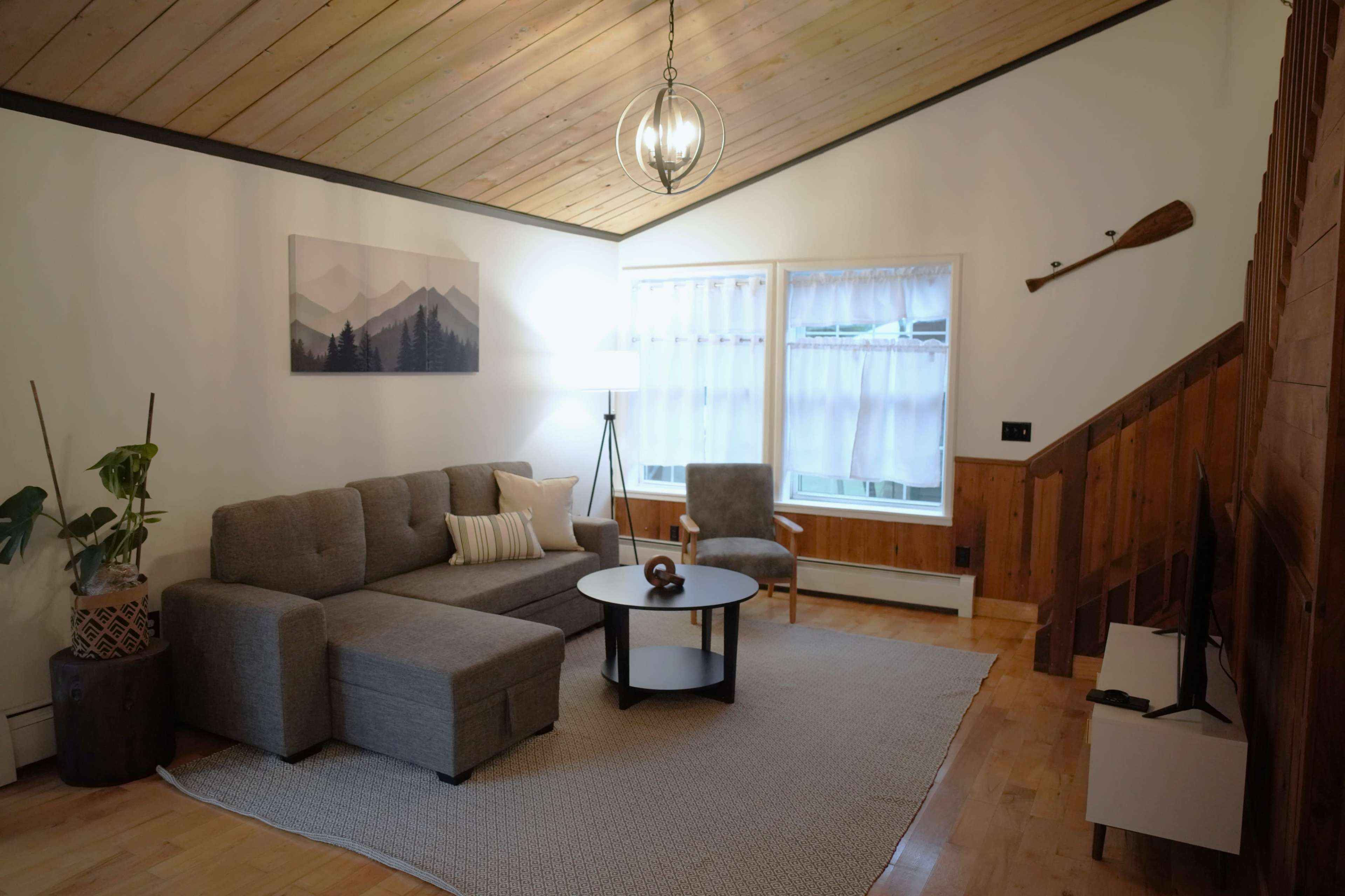 A cozy living room with a gray sectional sofa, a round coffee table, and large windows, featuring wooden accents and a high ceiling.