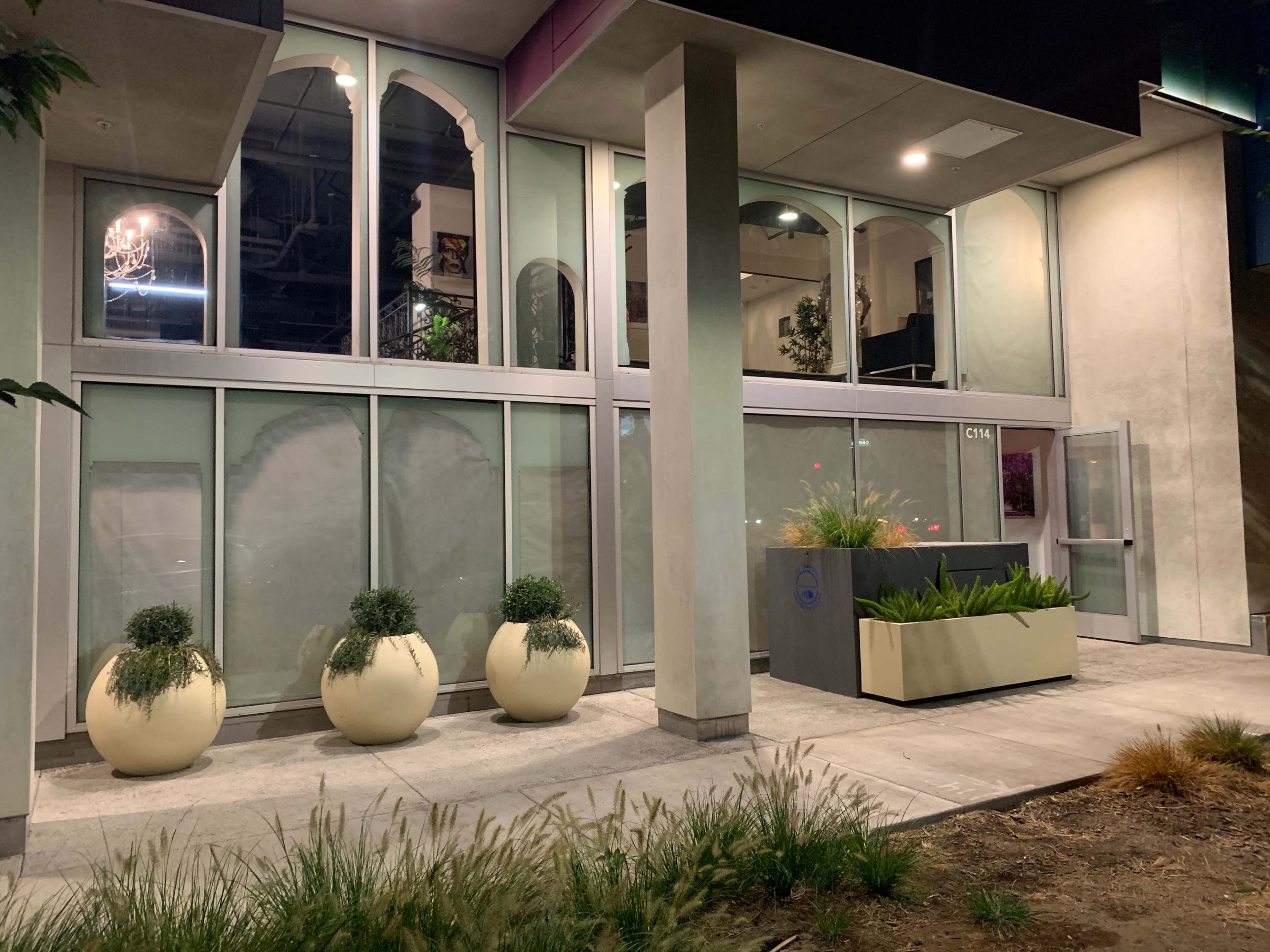 A modern building with large glass windows and planters near the entrance, illuminated at night.