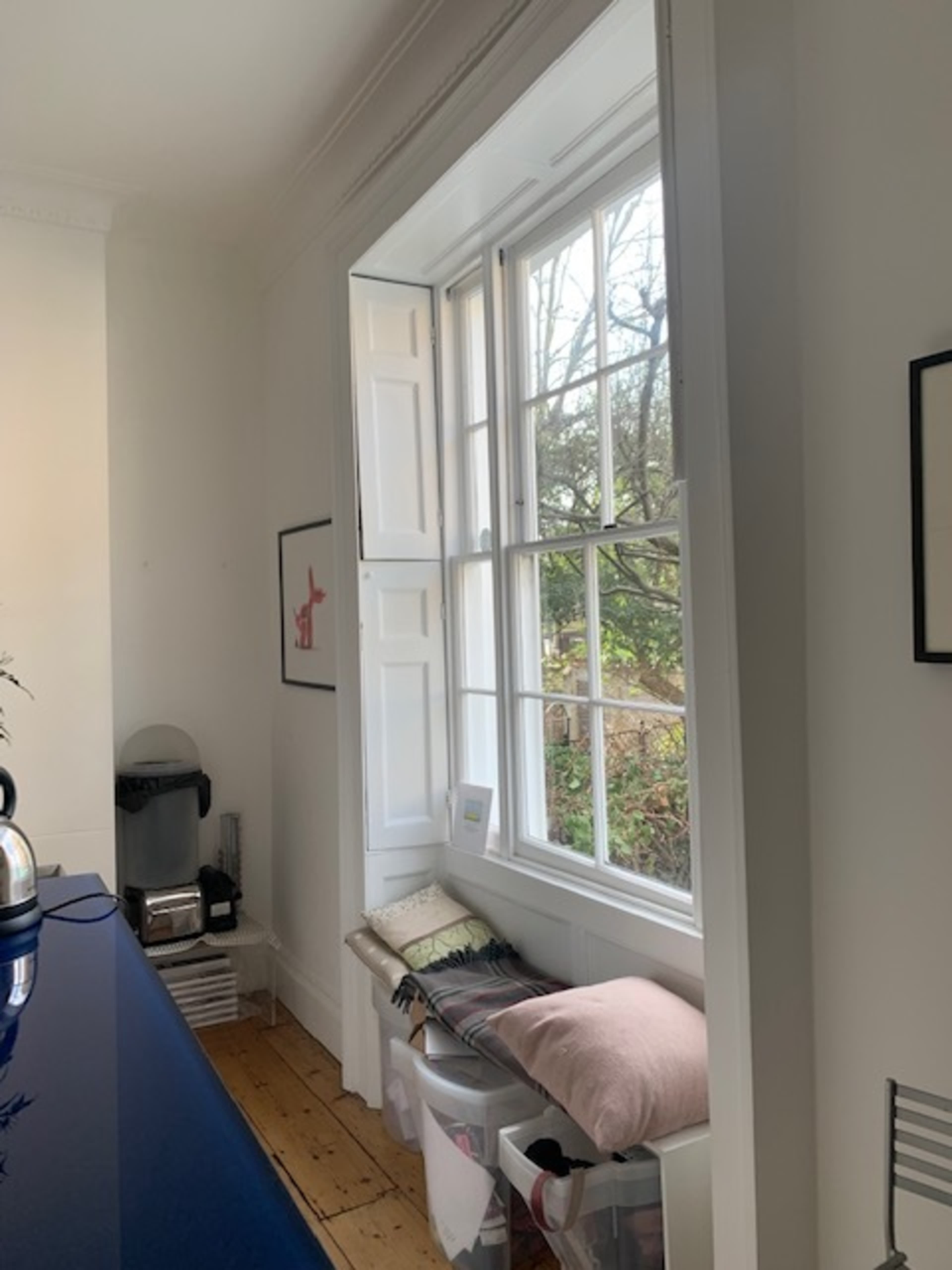An open window with white shutters, revealing a view of trees outside, next to a wall with framed art and a countertop with kitchen appliances.