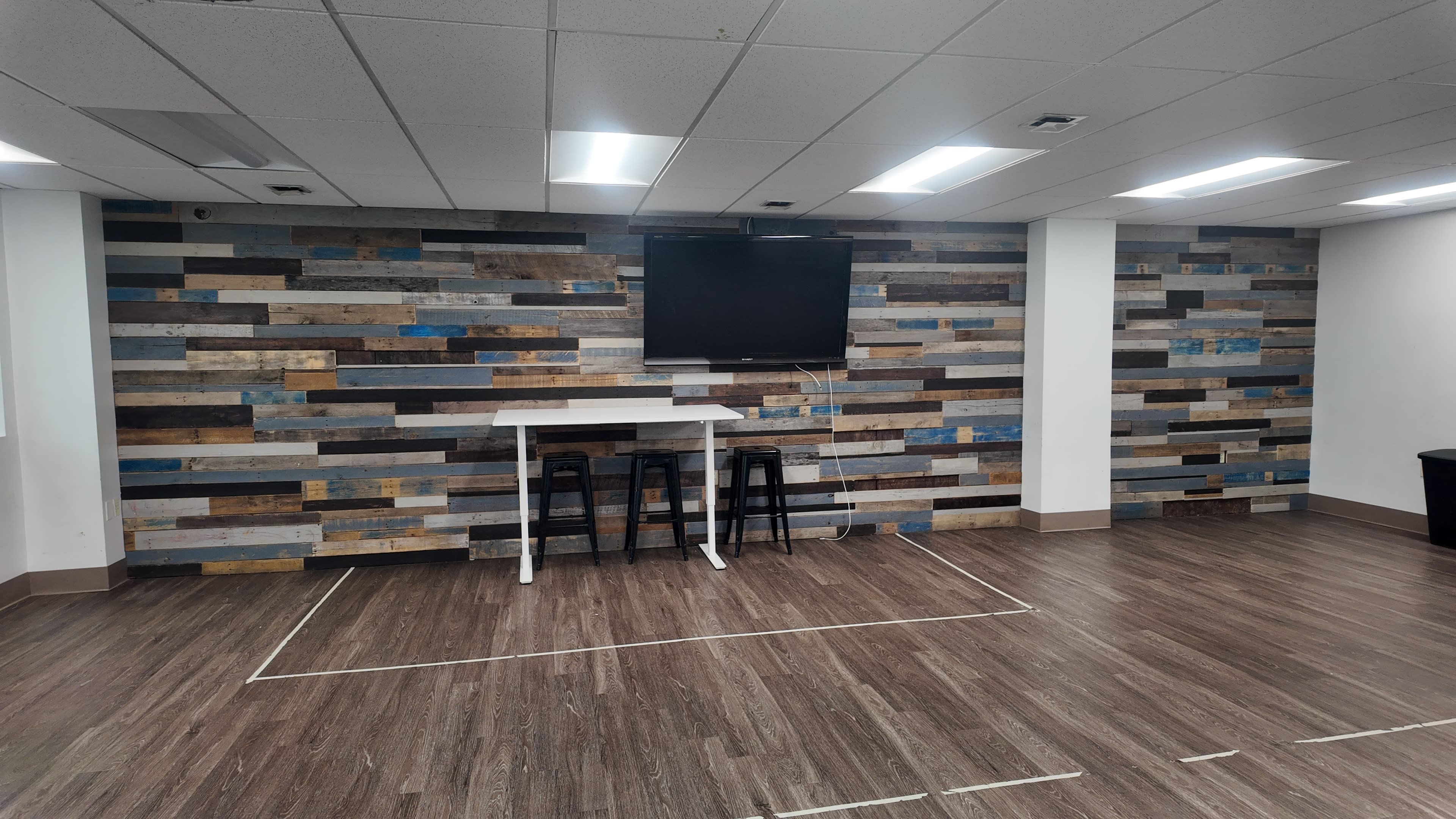 The image shows a modern indoor space featuring a textured wooden wall, a flat-screen television mounted above a white table, and two black stools.