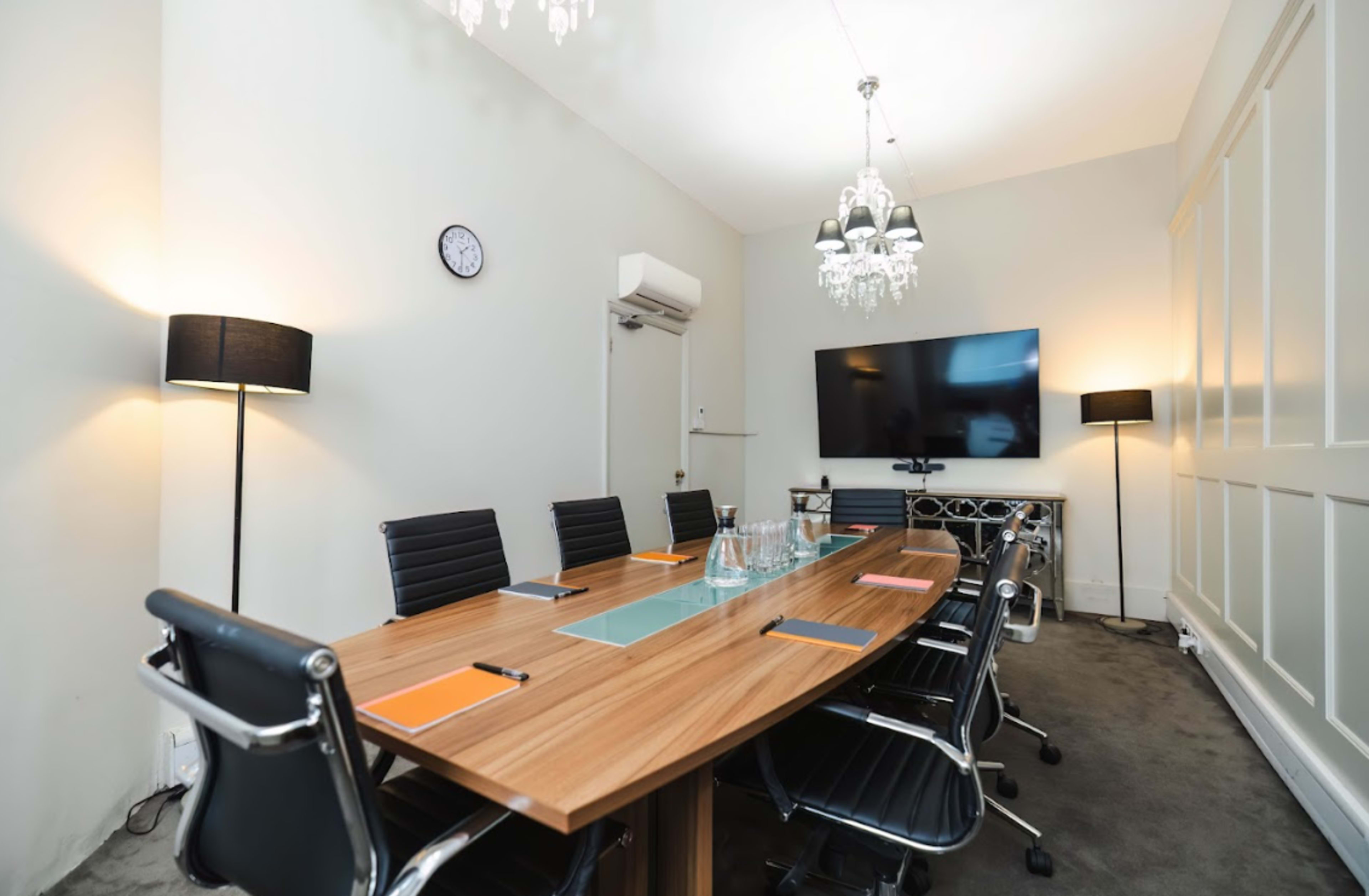 A modern conference room features a long wooden table surrounded by ergonomic chairs, with a large television and elegant lighting fixtures.