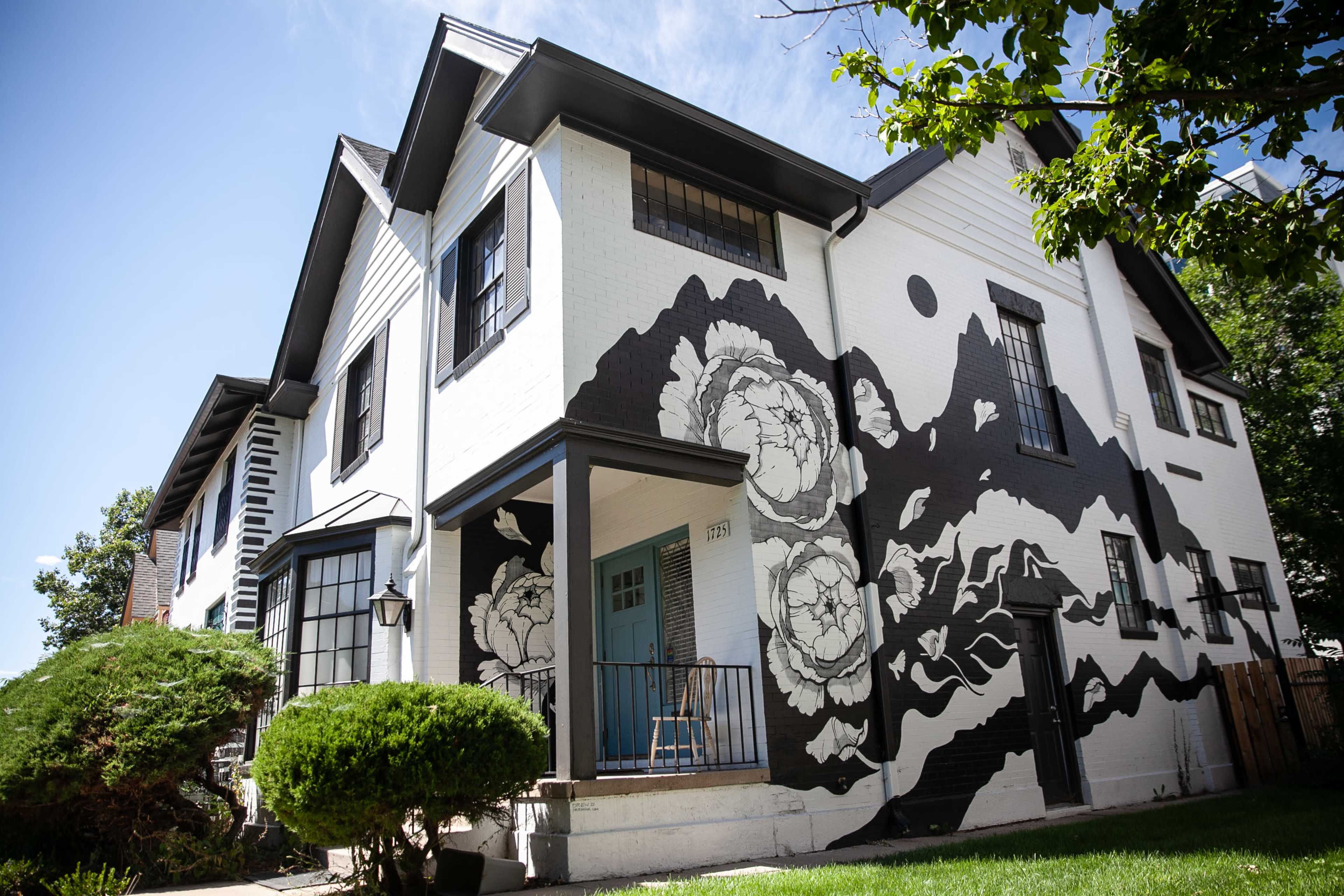 A house features a large black-and-white floral mural on one side, with a front porch and well-maintained landscaping.