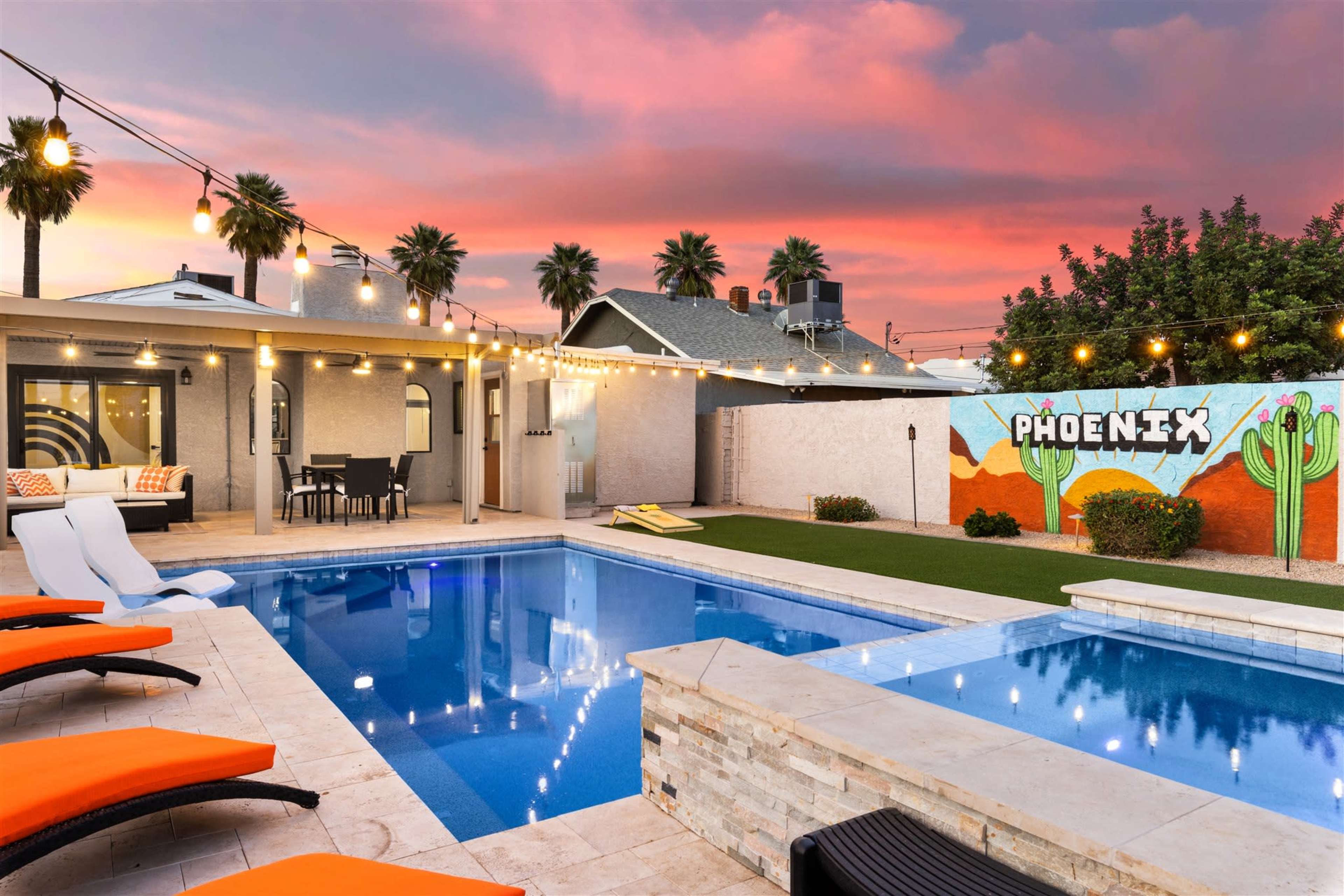 A swimming pool with lounge chairs is illuminated by string lights against a backdrop of a sunset sky and a mural reading "PHOENIX."