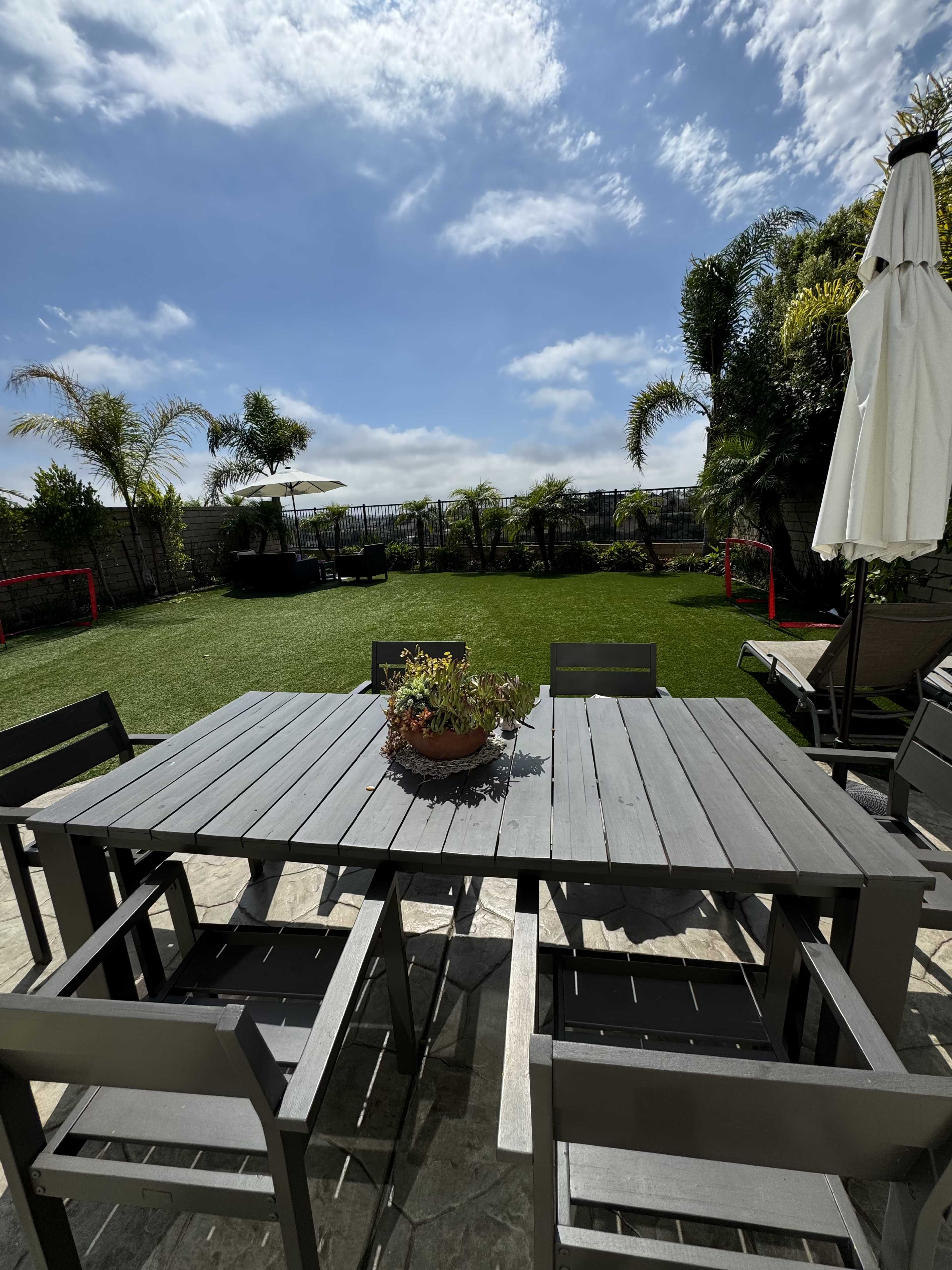 A gray outdoor dining table with chairs is set on a patio overlooking a landscaped yard with palm trees and a cloudy sky.